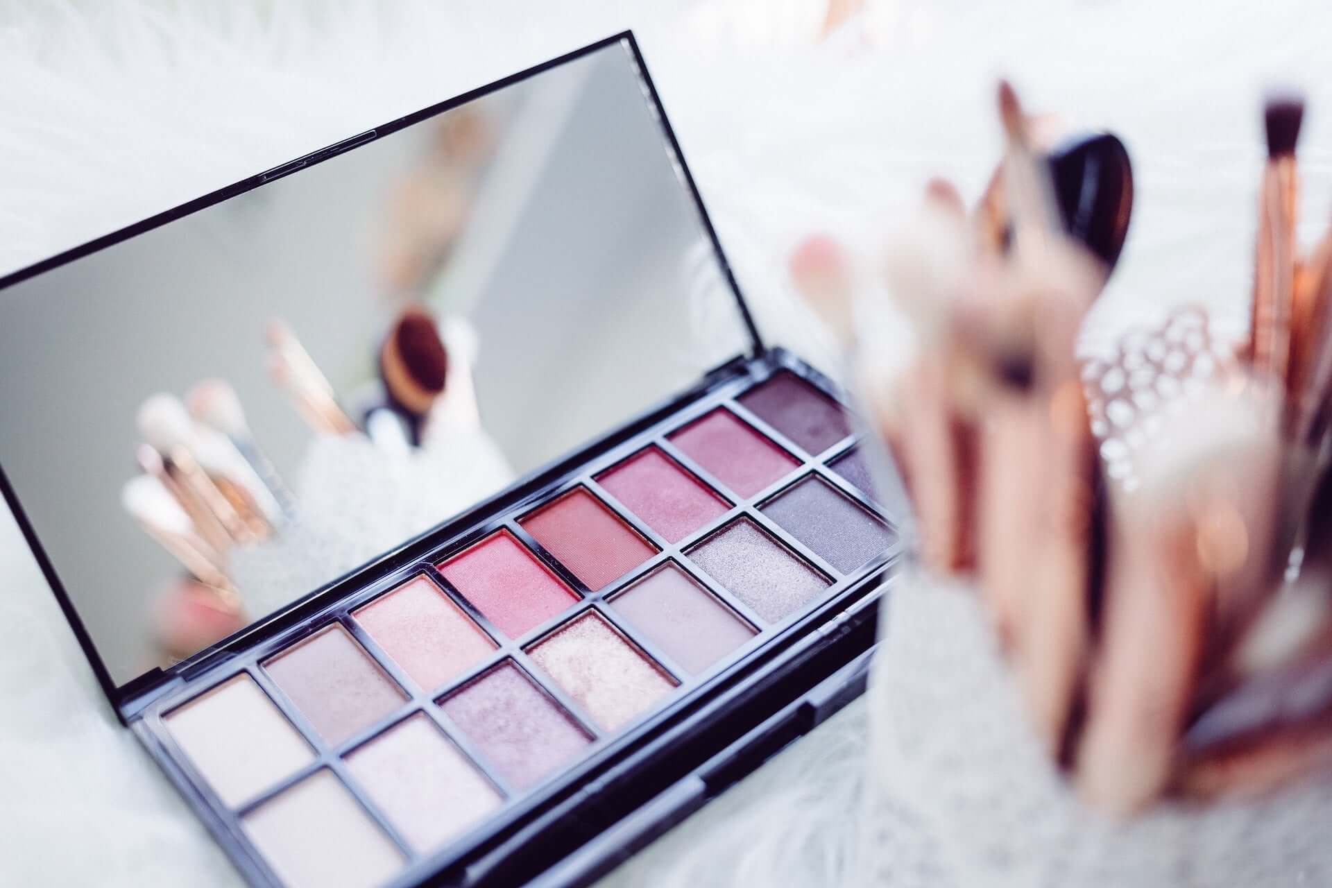 A pink-toned eyeshadow pallet with a mirror, sitting open beside a container of various cosmetic brushes.