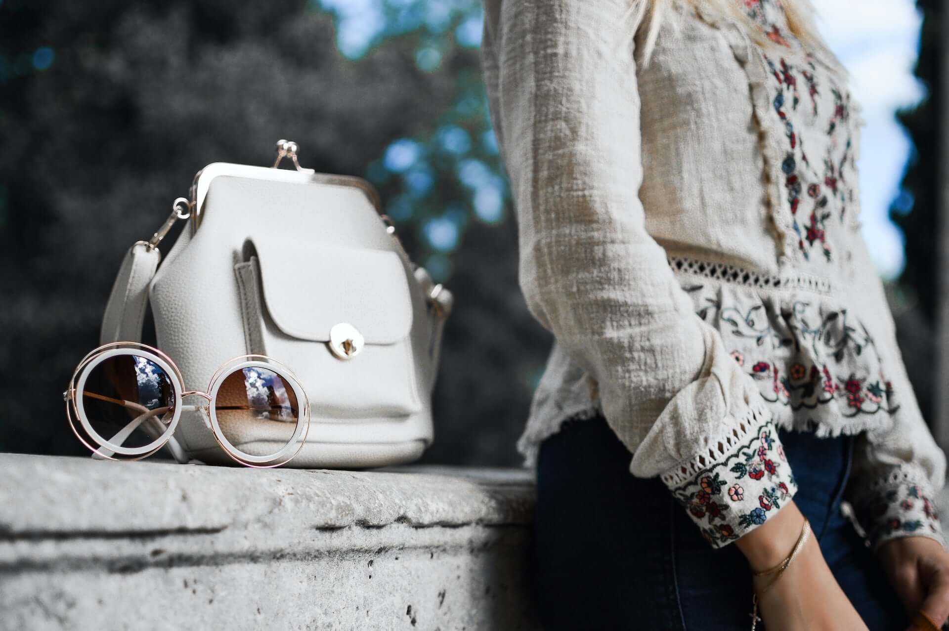 A woman in a flowing floral white top stands beside a white leather handbag and white round sunglasses.