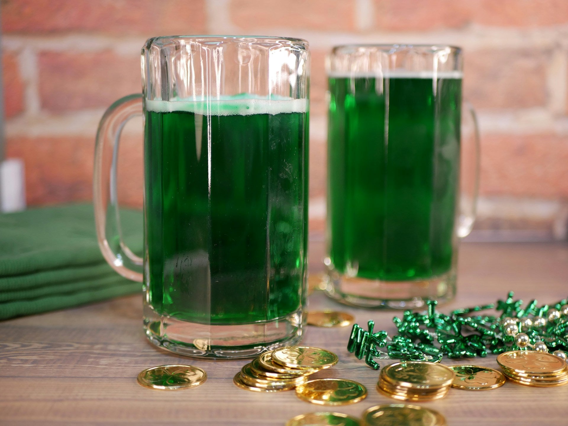 Two pints of green beer in glass mugs beside fake coins and green beads.