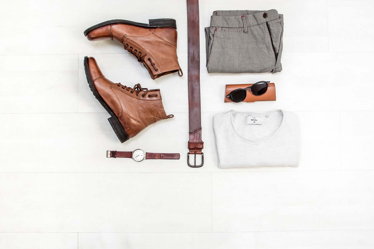 A flat overhead shot of an outfit incluing a pair of brown boots, a brown belt, a watch, sunglasses, a white shirt, and grey shorts.