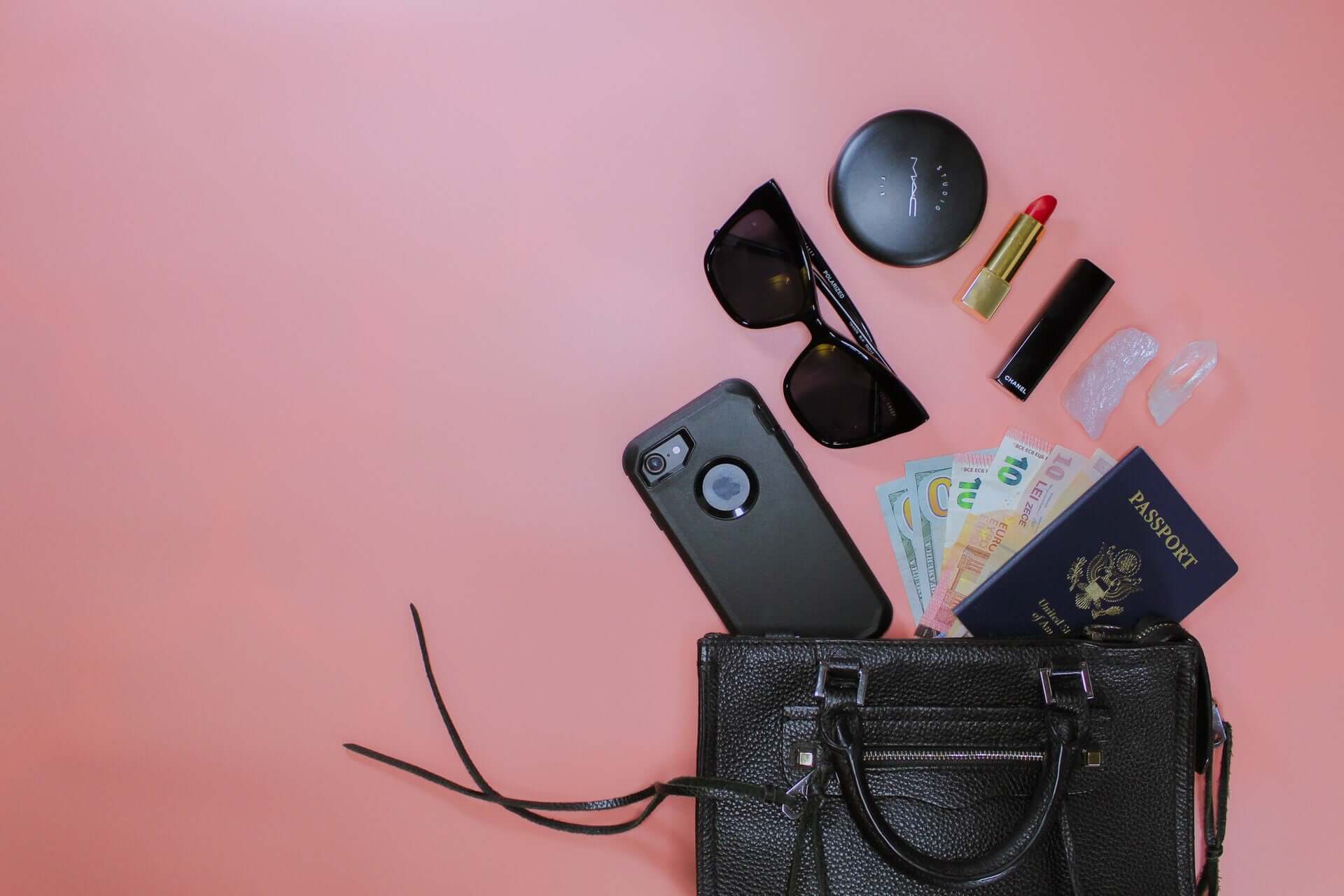 A black leather purse spills its contents - including sunglasses, a cellphone, a passport, and various makeup products - on a pink background.