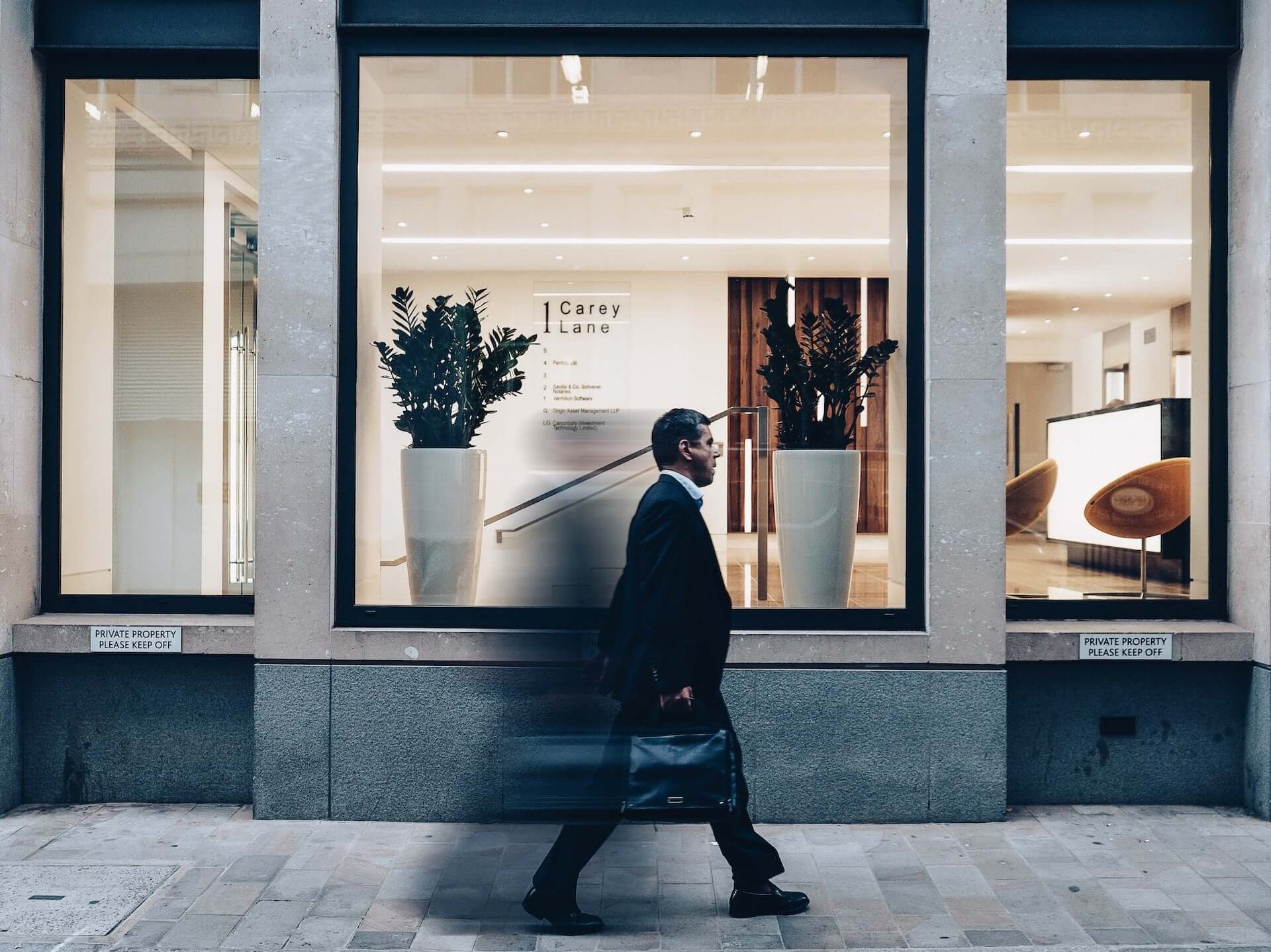 A man in a suit with a black briefcase walks by a large-windowed office.