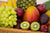 A basket containing grapes, mango, plums, apples, kiwi, pineapple, an other fruits.