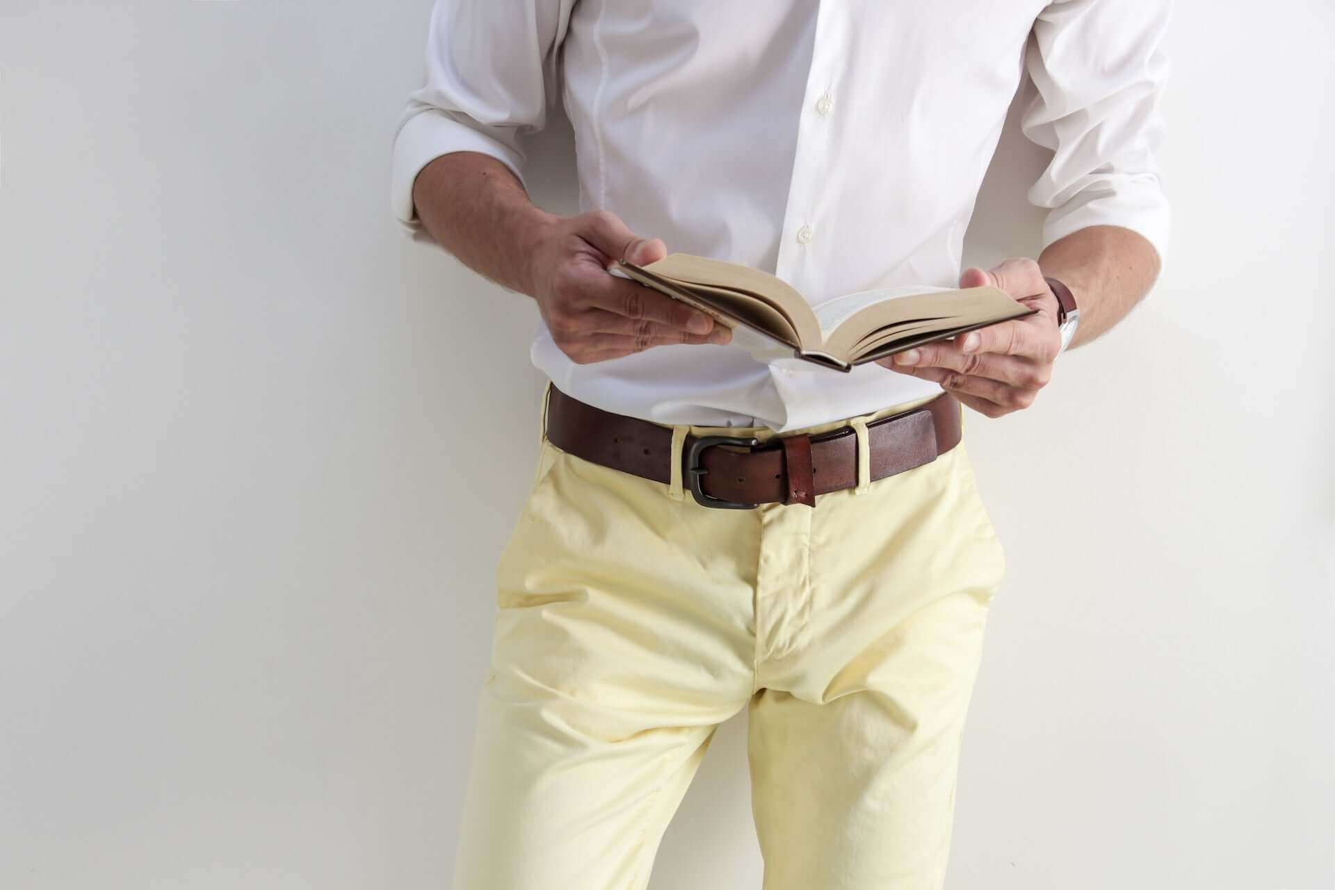 A man in a white button down, yellow slacks, and a brown leather belt. He holds a book.