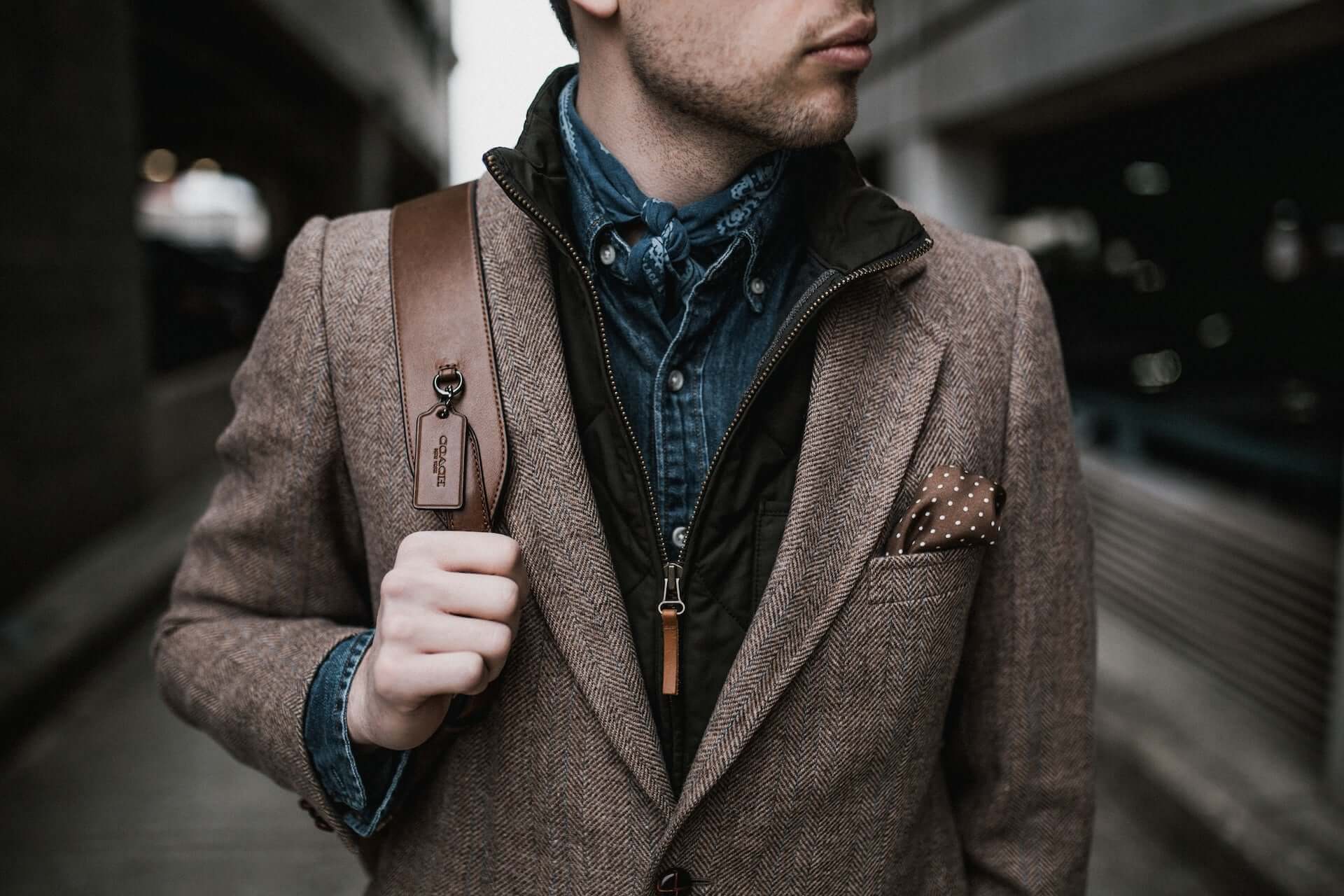 A man in a brown tweed blazer, zip-up grey jacket, and blue button down, carrying a Coach bag.