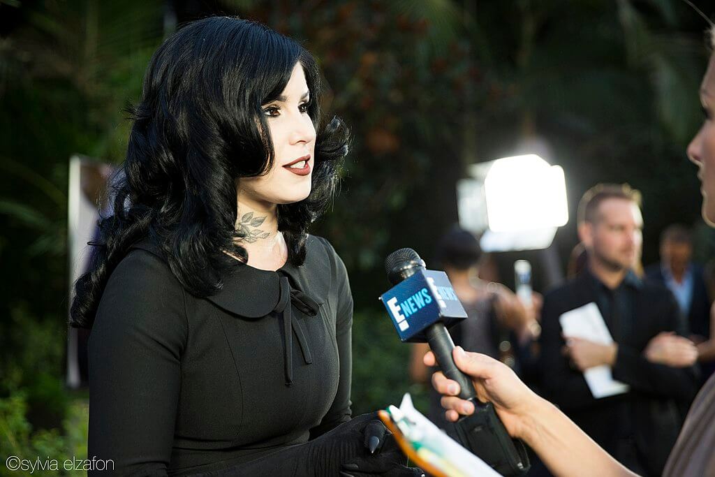 Kat Von D being interviewed. She is a white woman with black hair and tattoos, dressed all in black.