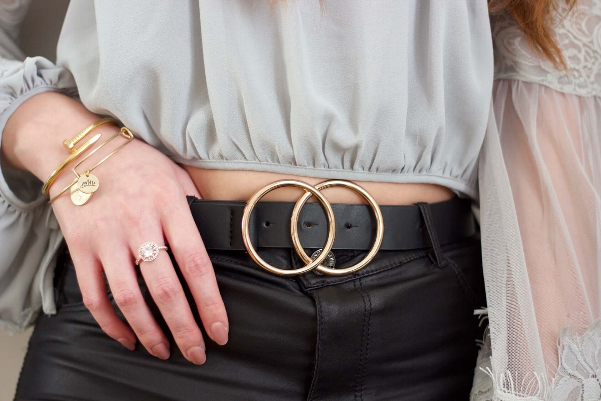 A close shot of a woman's midsection. She wears a grey blouse and black jeans, and has a black belt with a buckle composed of two linked circles.