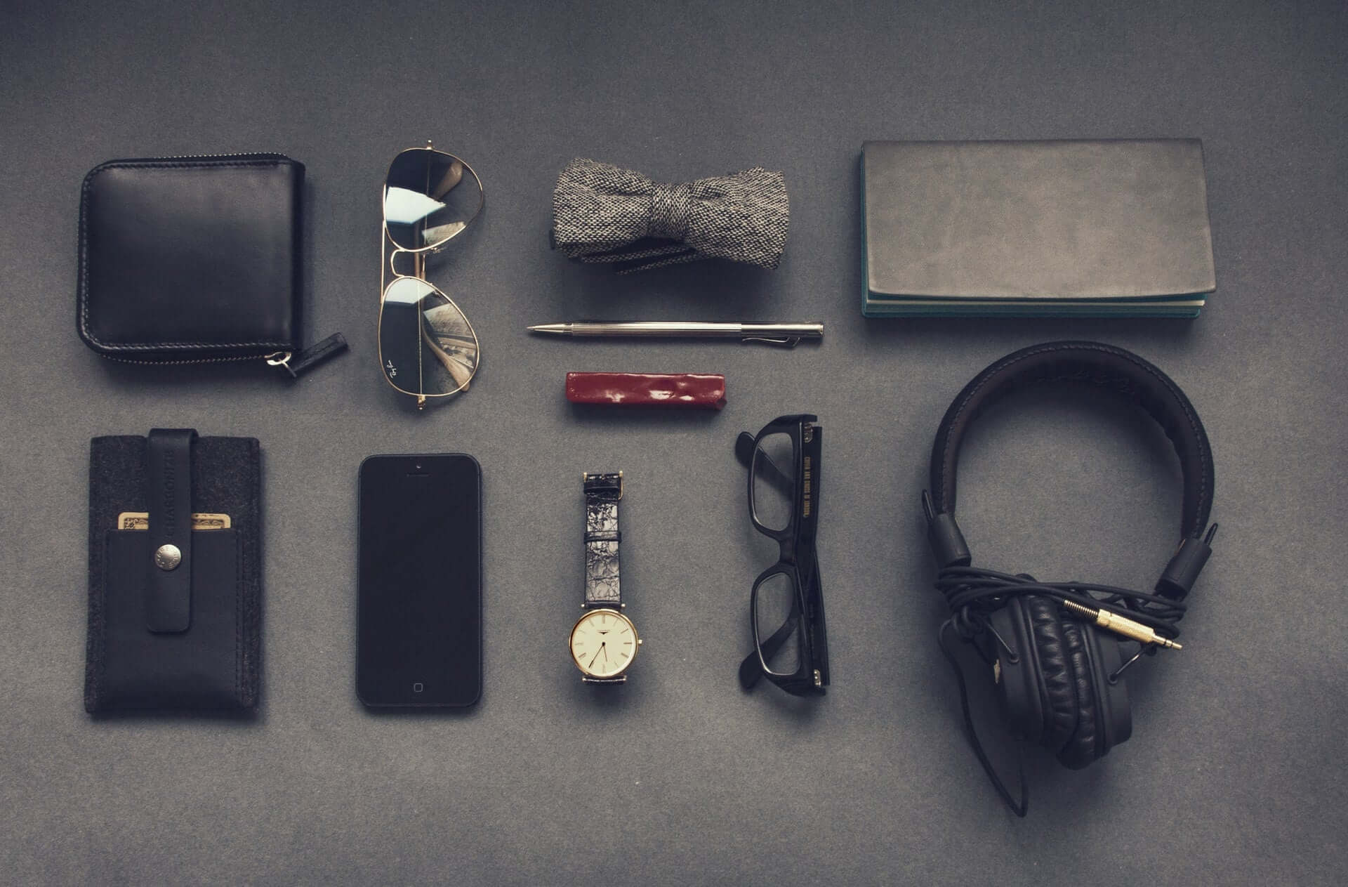 An overhead shot of a flatlay of day-to-day items including a wallet, sunglasses, glasses, headphones, a watch, a phone, and a bowtie.