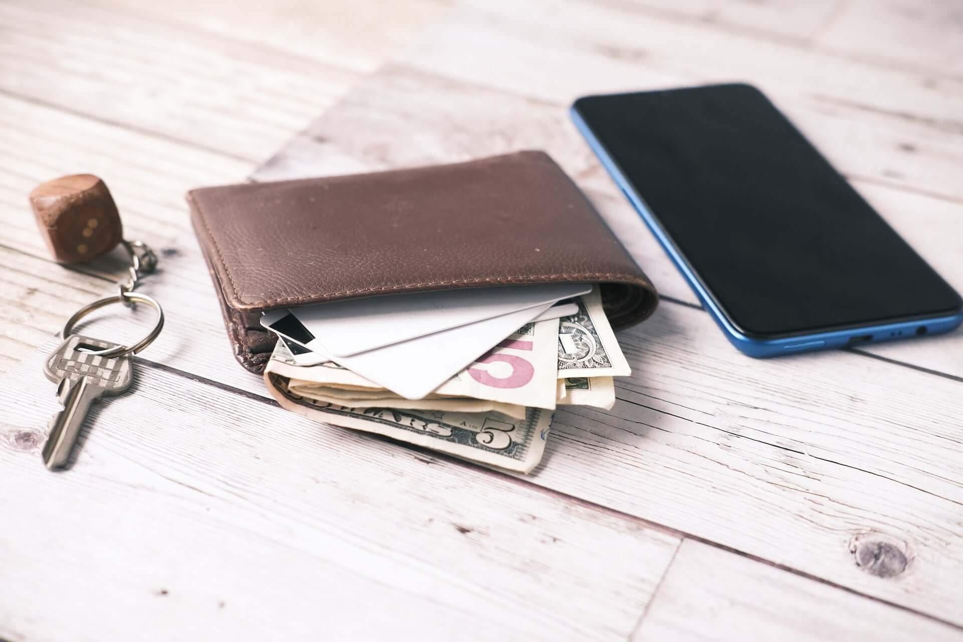A bifold wallet full of American dollar bills sits beside a smartphone and a set of keys.