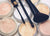 A mascara wand, eyeshadow brush, and angled blush brush sit on various cosmetic powders.
