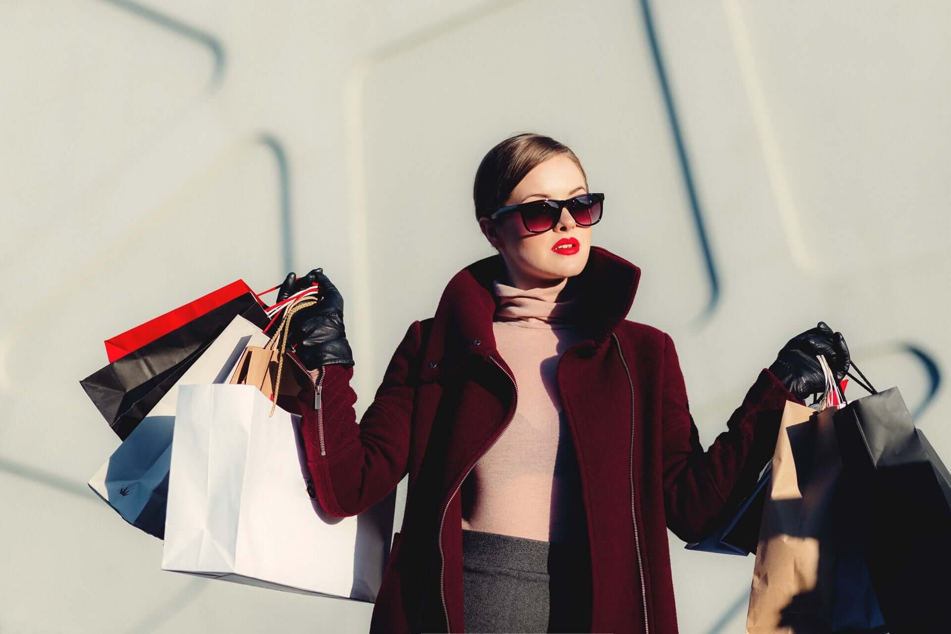 A woman in a burgandy coat, pink sweater, and sunglasses holds a variety of shopping bags as she poses.