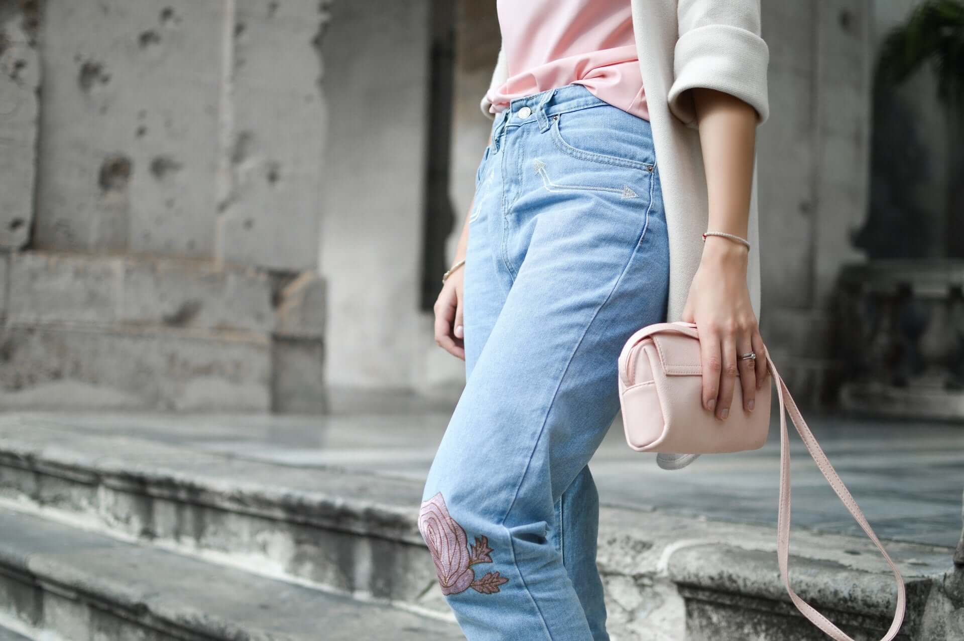 A woman in jeans, a pink top, and a cream sweater holds a pink clutch bag to one side.