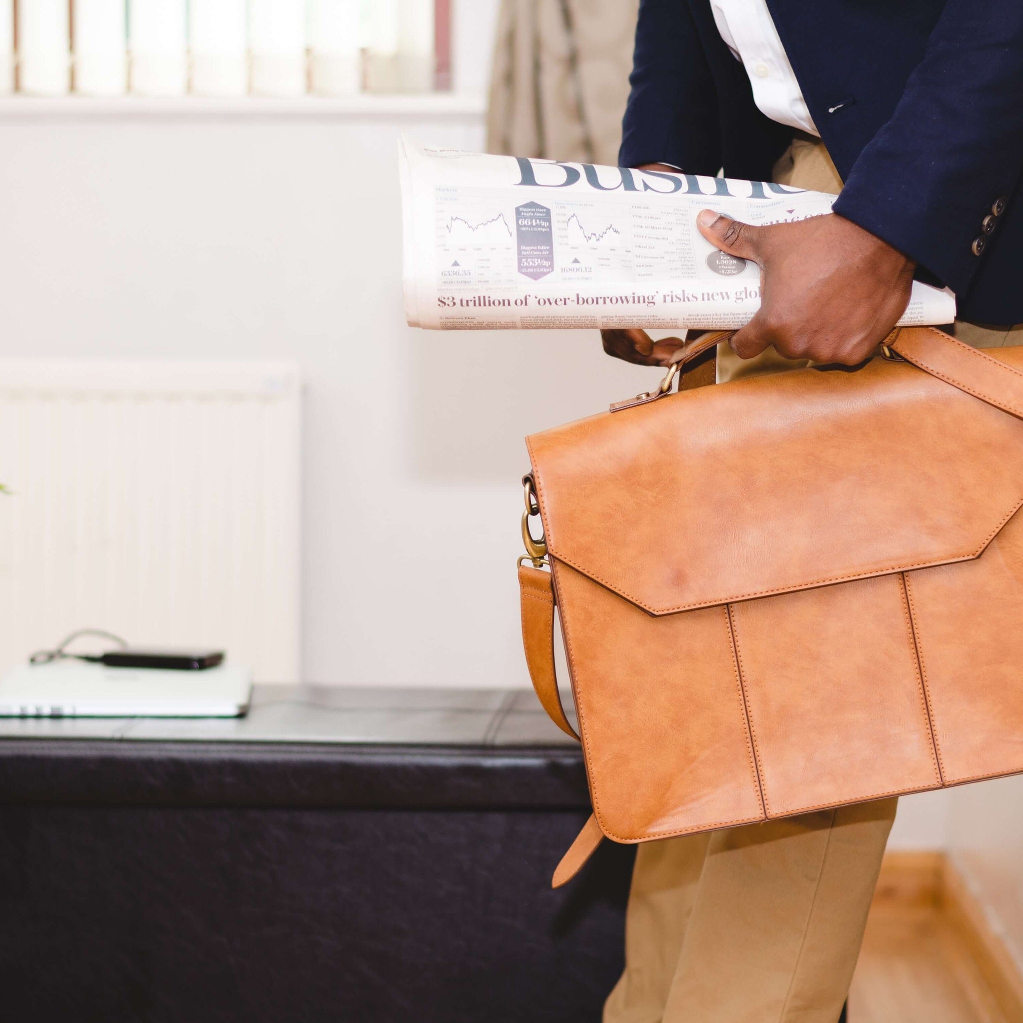 A cutoff image of a man in business attire holding a tan briefcase and a newspaper.