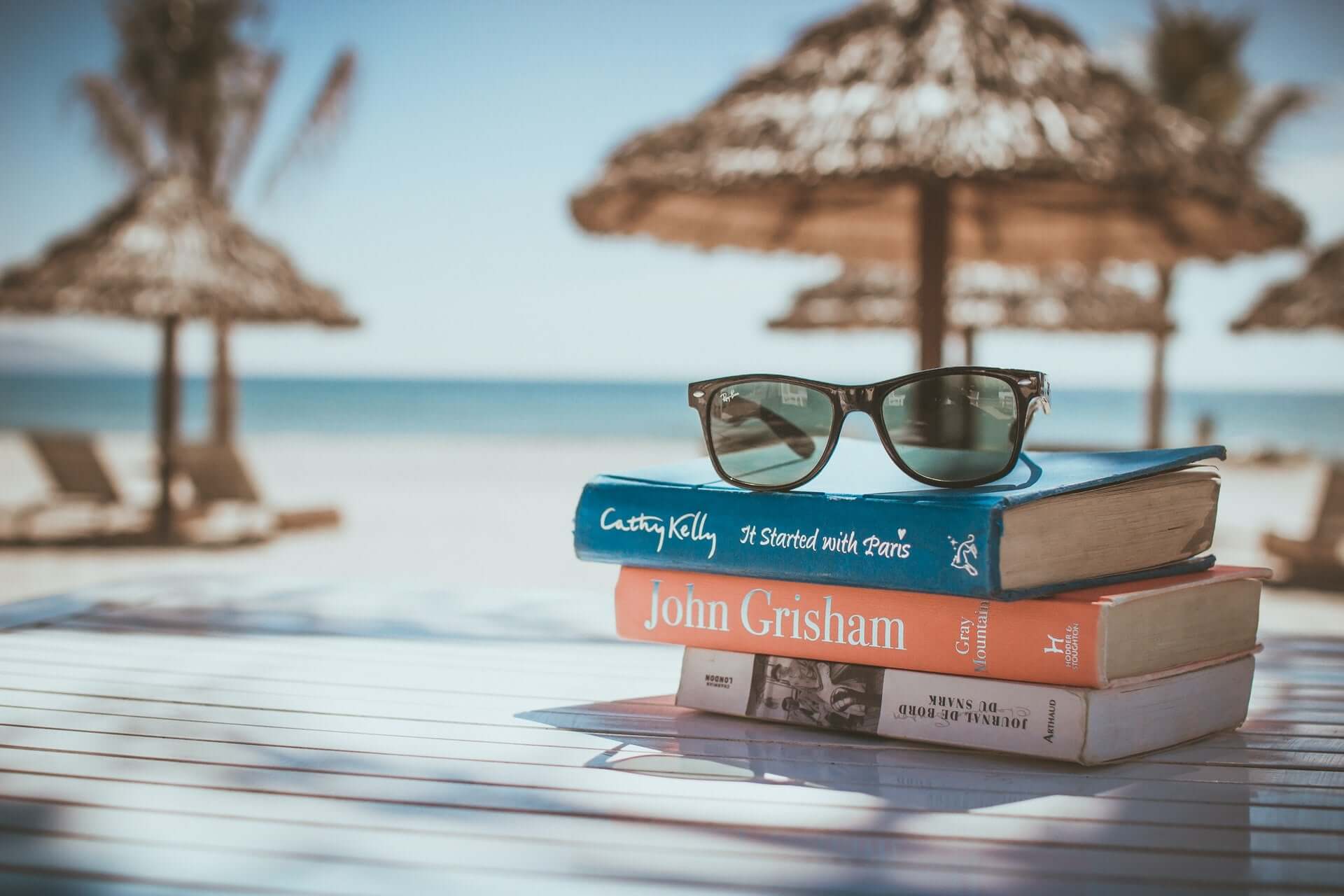 A stack of books and a pair of sunglasses on a table on a white sand beach.