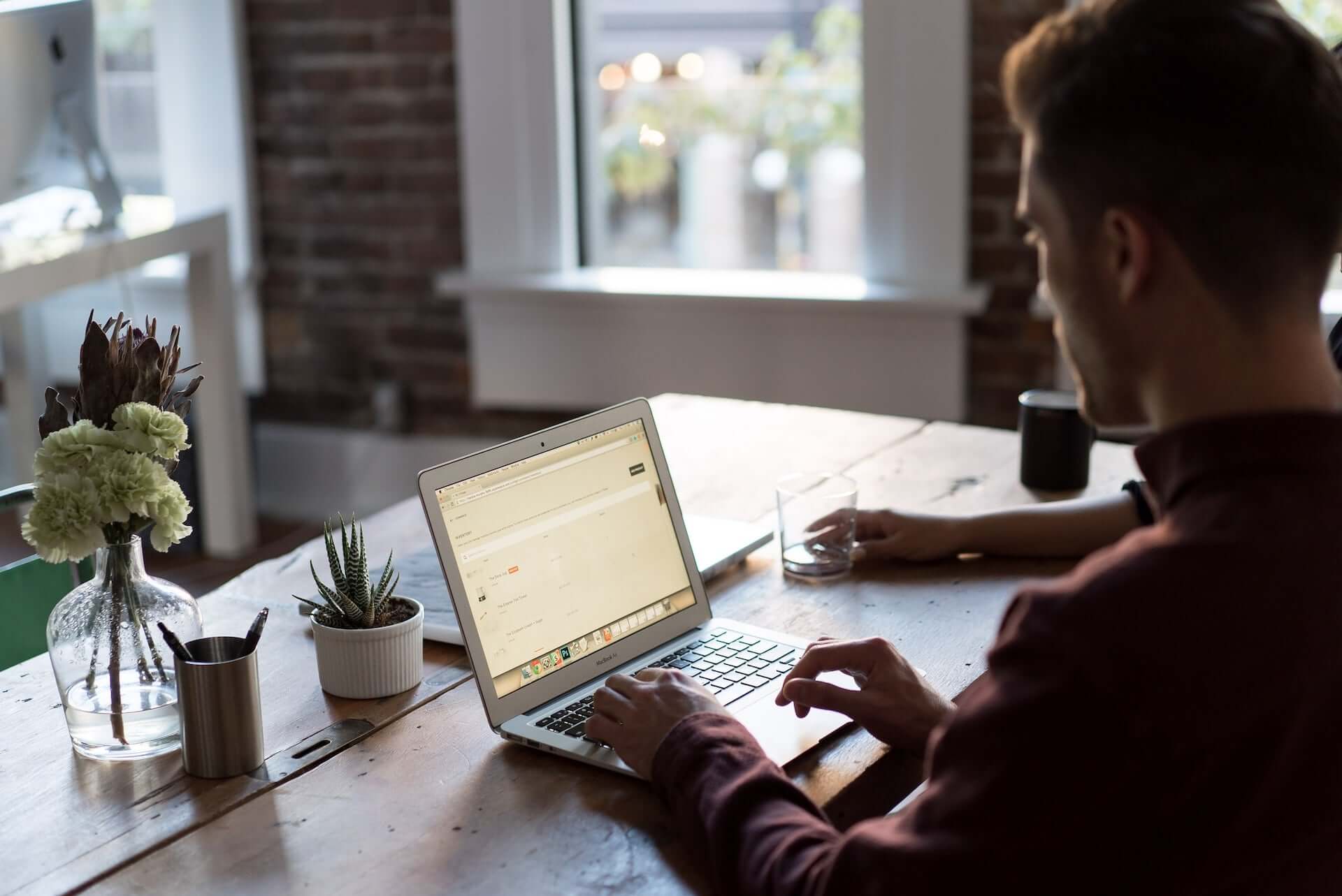 A man types on a laptop while sitting at a wooden table in a brick office.