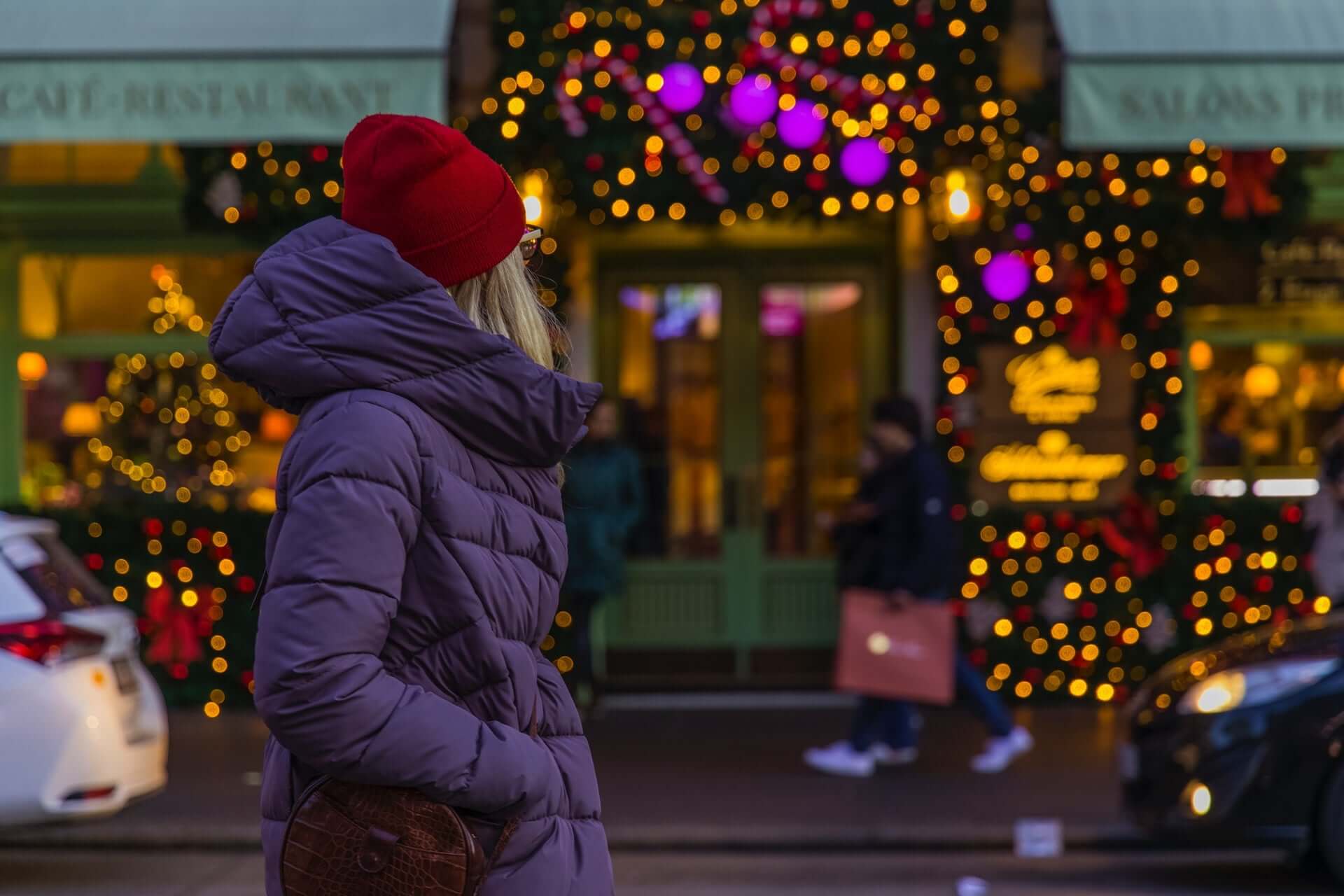 A woman in a black puffer jacket and red beanie stands facing away from the camera in front of a store decorated with lights.