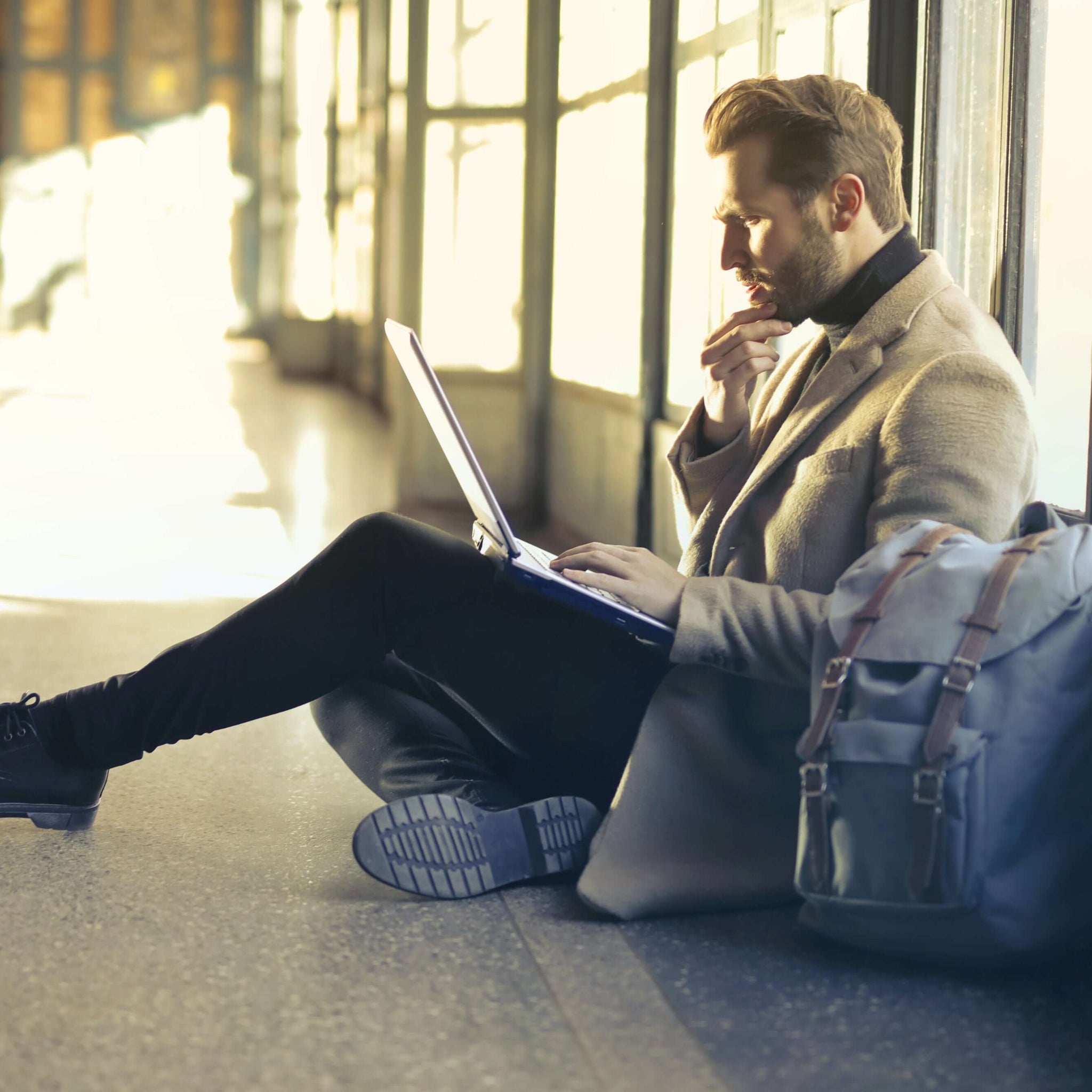 A man in a brown suit jacket and slacks is seated in front of a tall floor-to-ceiling window, working on a laptop.