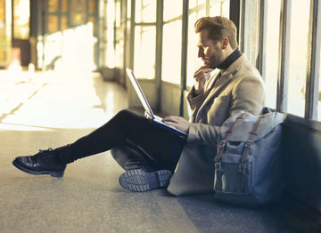 A man in a brown suit jacket and slacks is seated in front of a tall floor-to-ceiling window, working on a laptop.