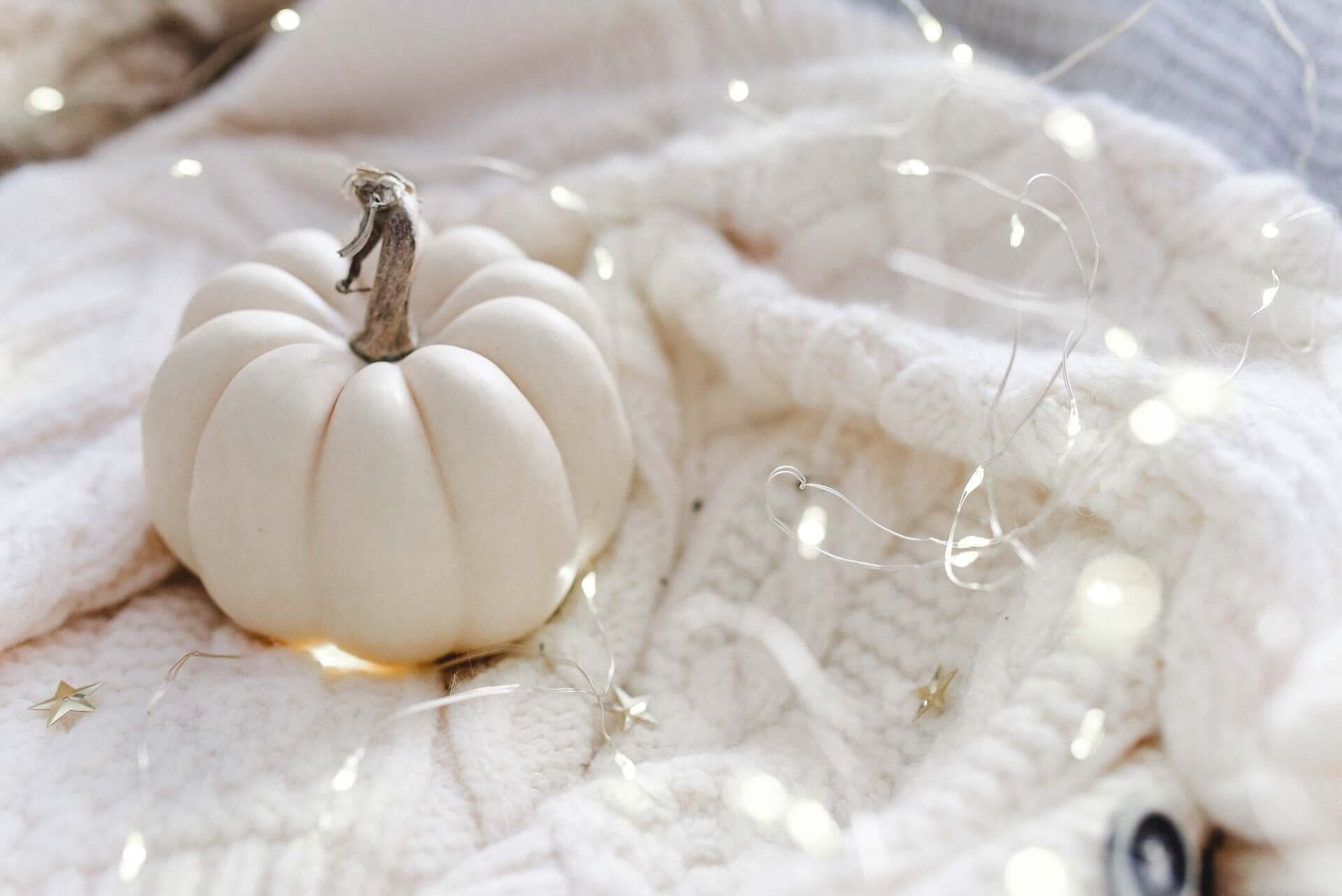 A white pumpkin sitting on a white knit blanket covered in fairy lights.