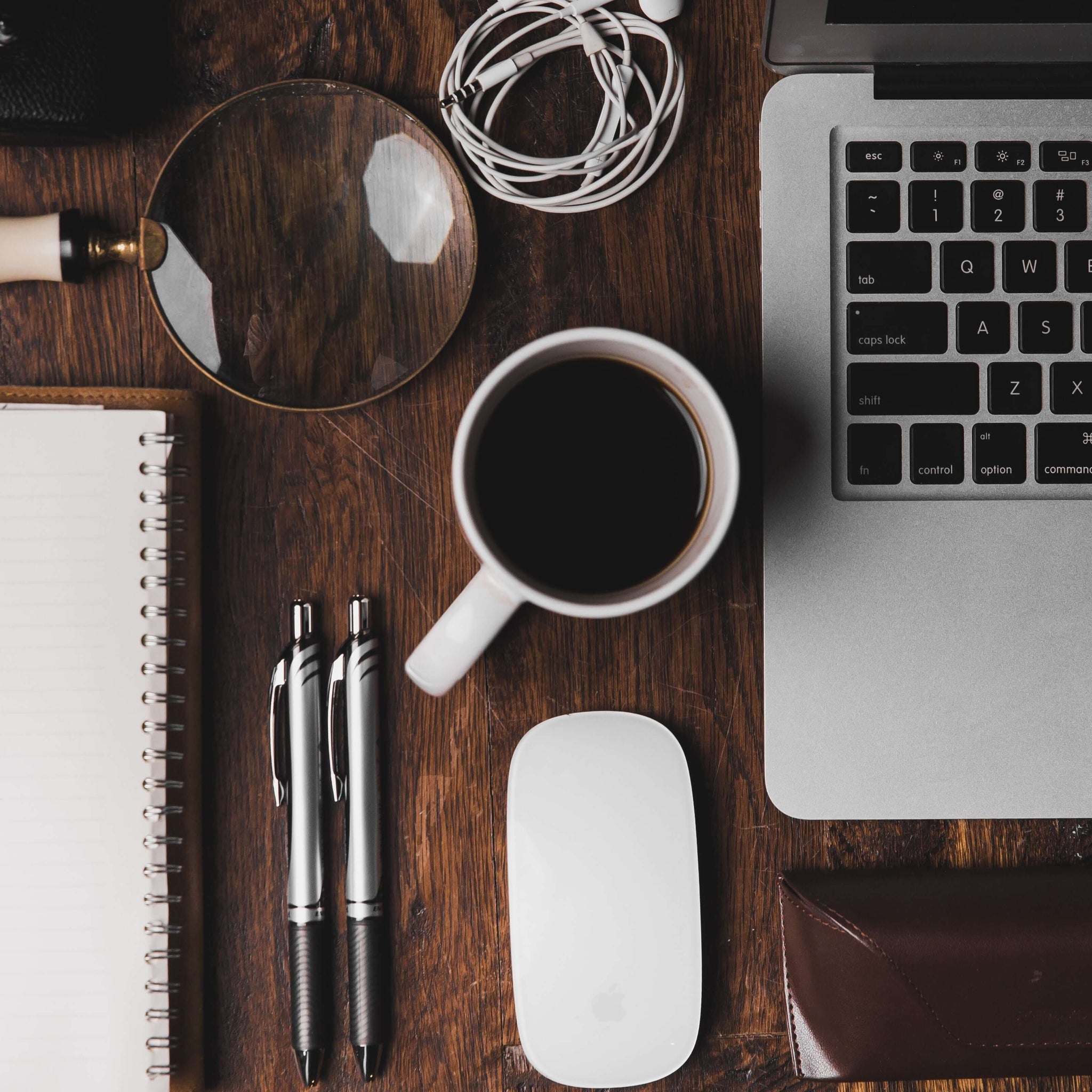 An overead shot of a desk with well-organized supplies including a laptop, two pens, an open notebook, and curled headphones. A cup of coffee is in the center of the spread.