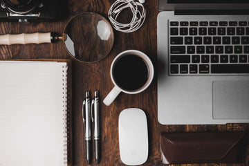 An overead shot of a desk with well-organized supplies including a laptop, two pens, an open notebook, and curled headphones. A cup of coffee is in the center of the spread.