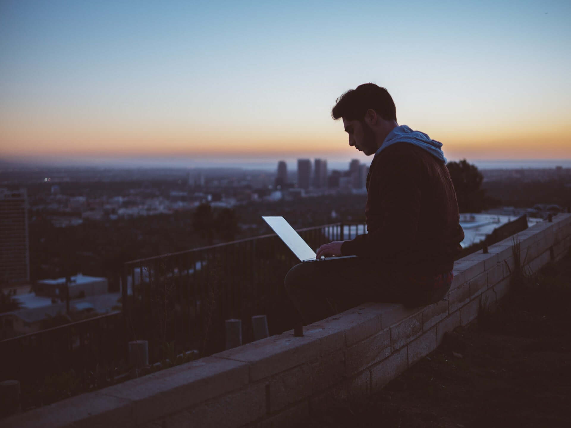 A man in a hoodie sits on the edge of a city roof, working on a laptop.