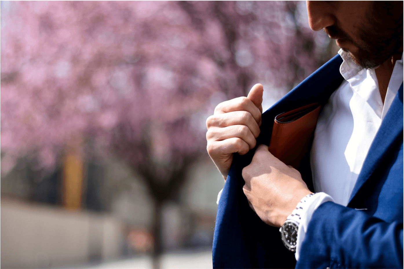 A man in a blue suit puts something away in an interior jacket pocket. He stands in front of a cherry blossom tree.