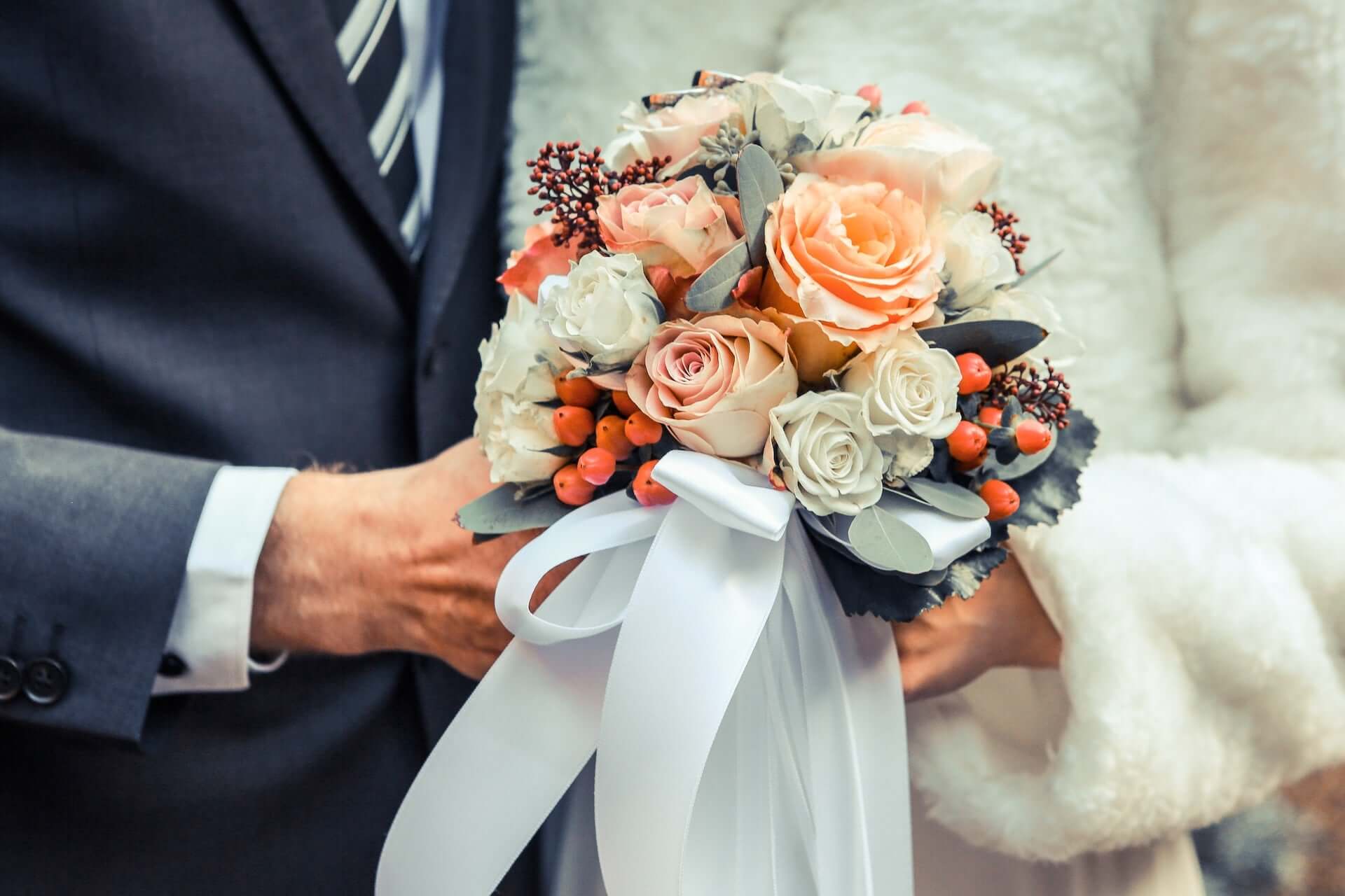 A wedding couple holding a bouquet of peach and white roses and holly berries.
