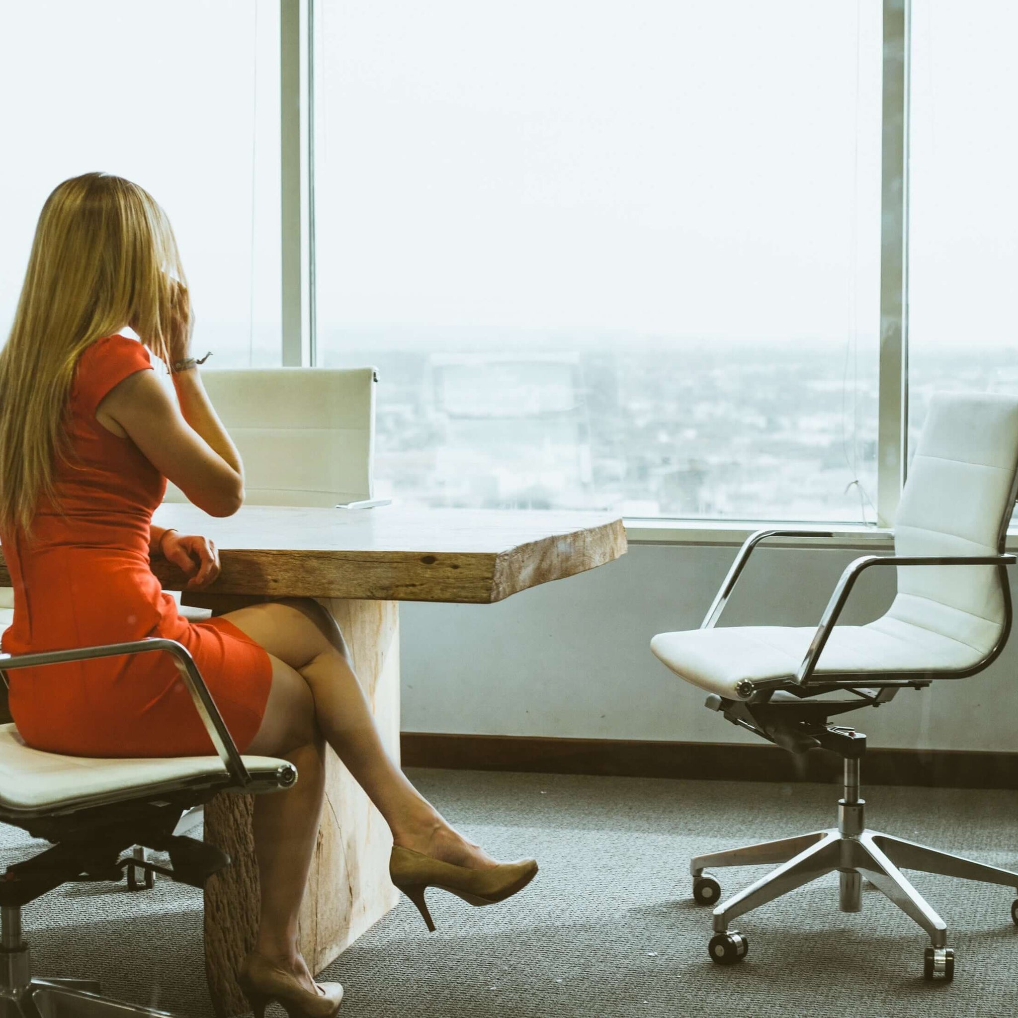 A woman in an orange dress sits in an office with white chairs and tables, and windows overlooking a hilly city.