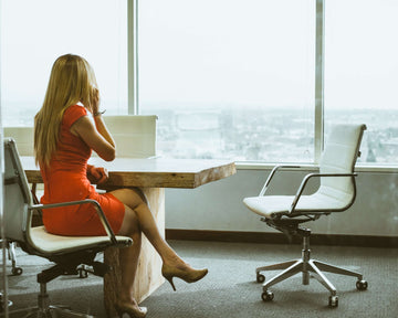 A woman in an orange dress sits in an office with white chairs and tables, and windows overlooking a hilly city.