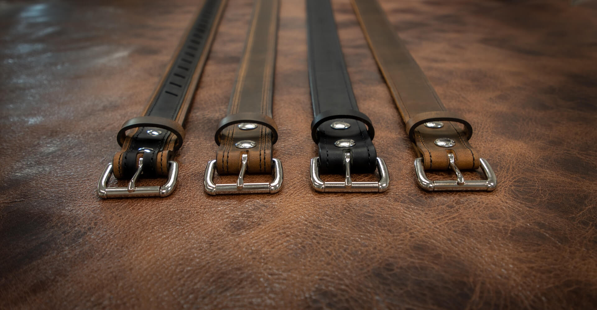 Four leather belts in various shades of black and brown laid flat on a brown surface.