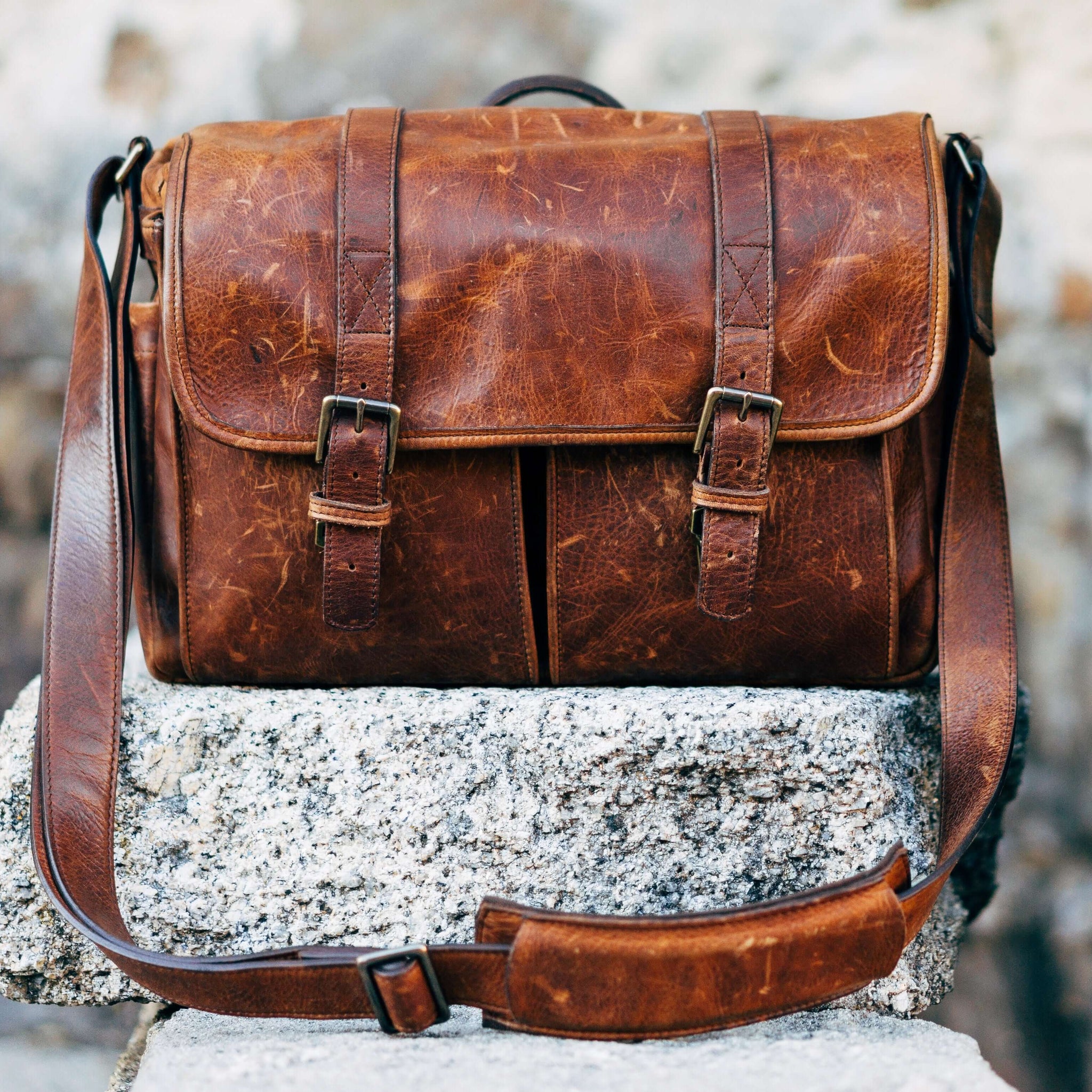 A brown messenger bag sitting on a grey stone, in front of a grey stone wall.