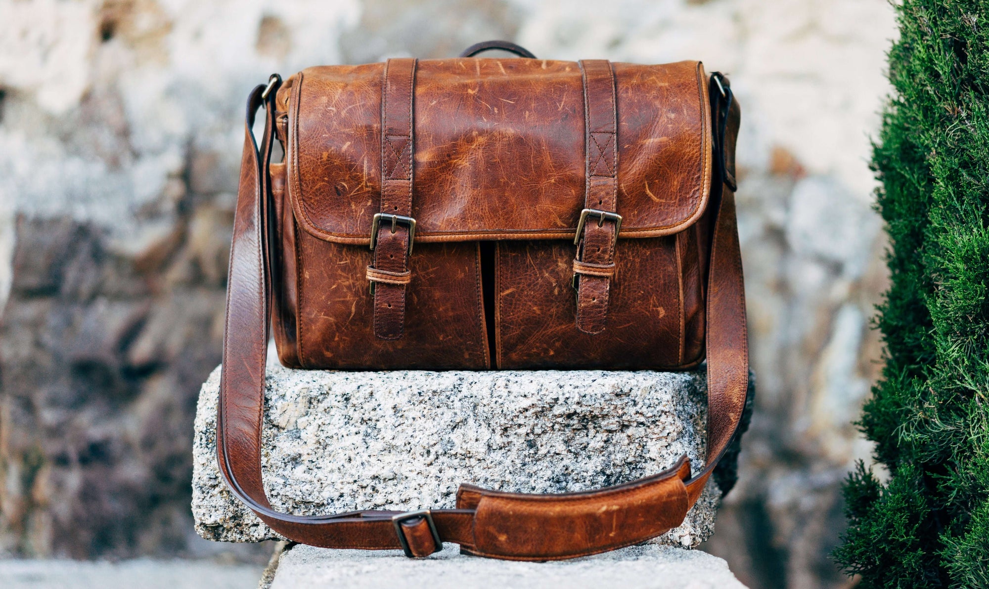 A brown messenger bag sitting on a grey stone, in front of a grey stone wall.