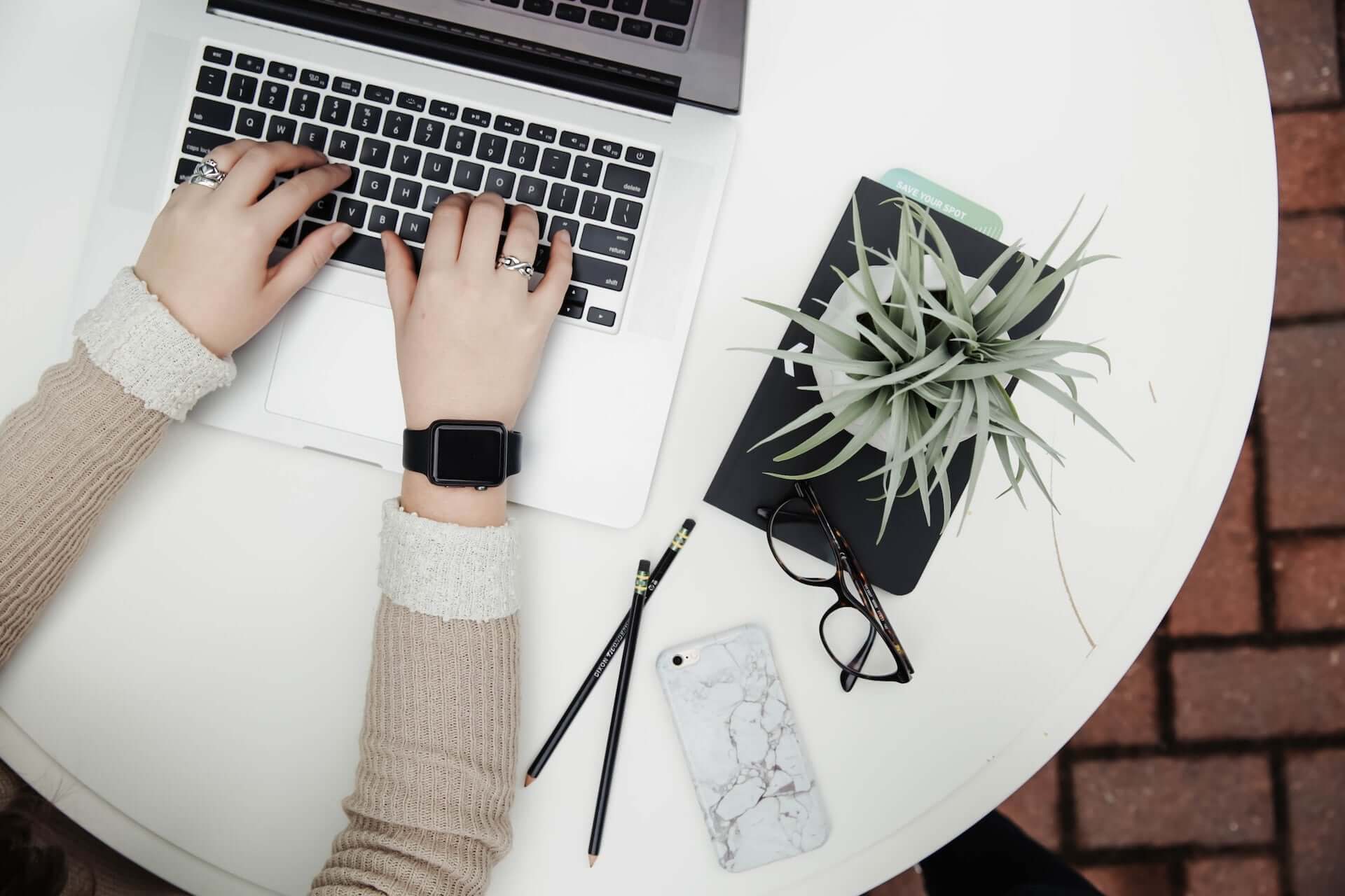 A person with painted nails and rings writes at a silver laptop. Beside them is a potted plant, glasses, pencils, and a phone.