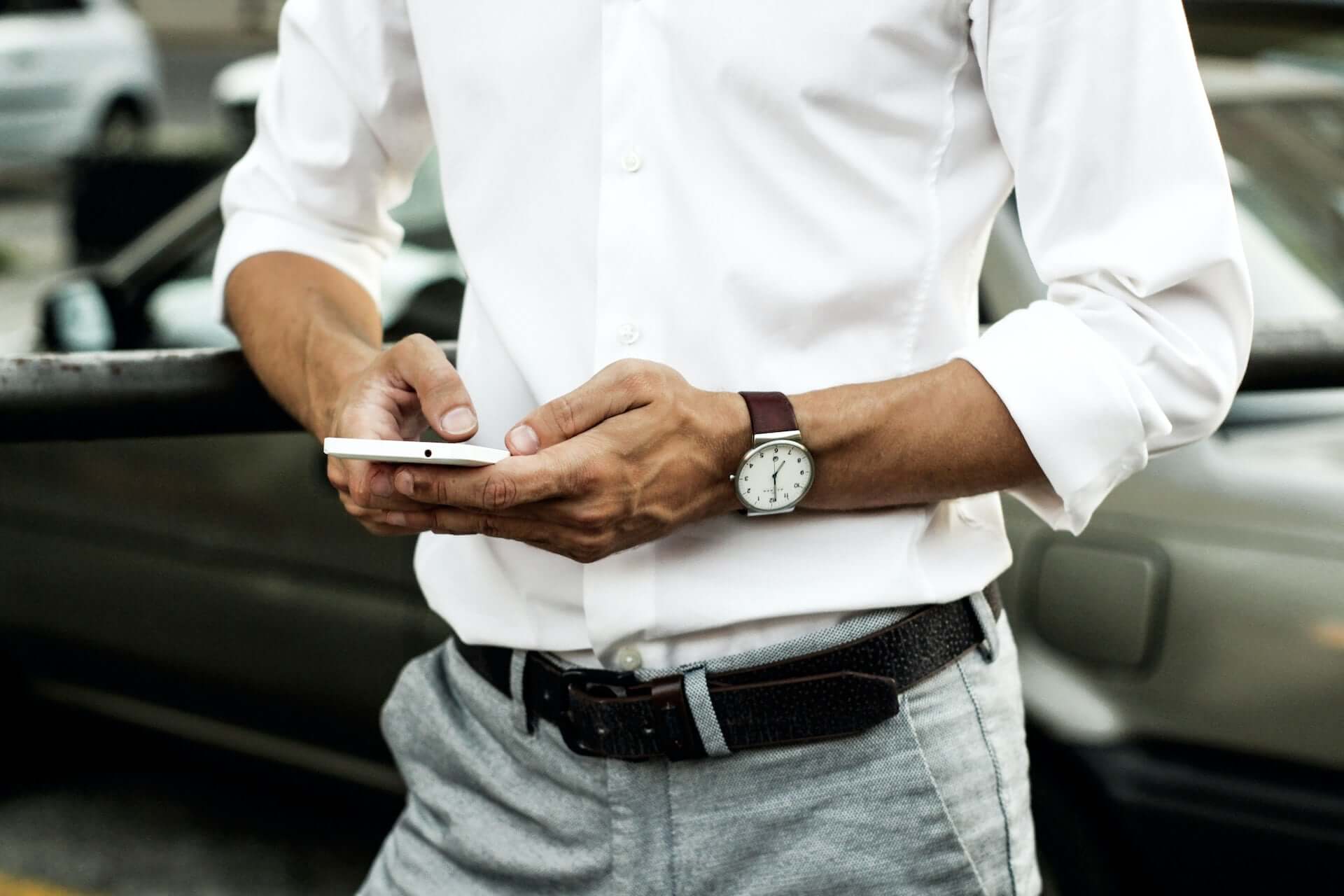 A man in a white shirt and light pants with a black belt types on a phone.