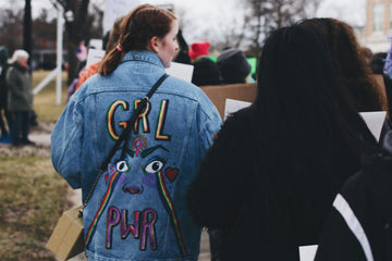 Two women walking away from the camera. One wears a denim jacket that reads "GRL PWR," with a supportive illustration.