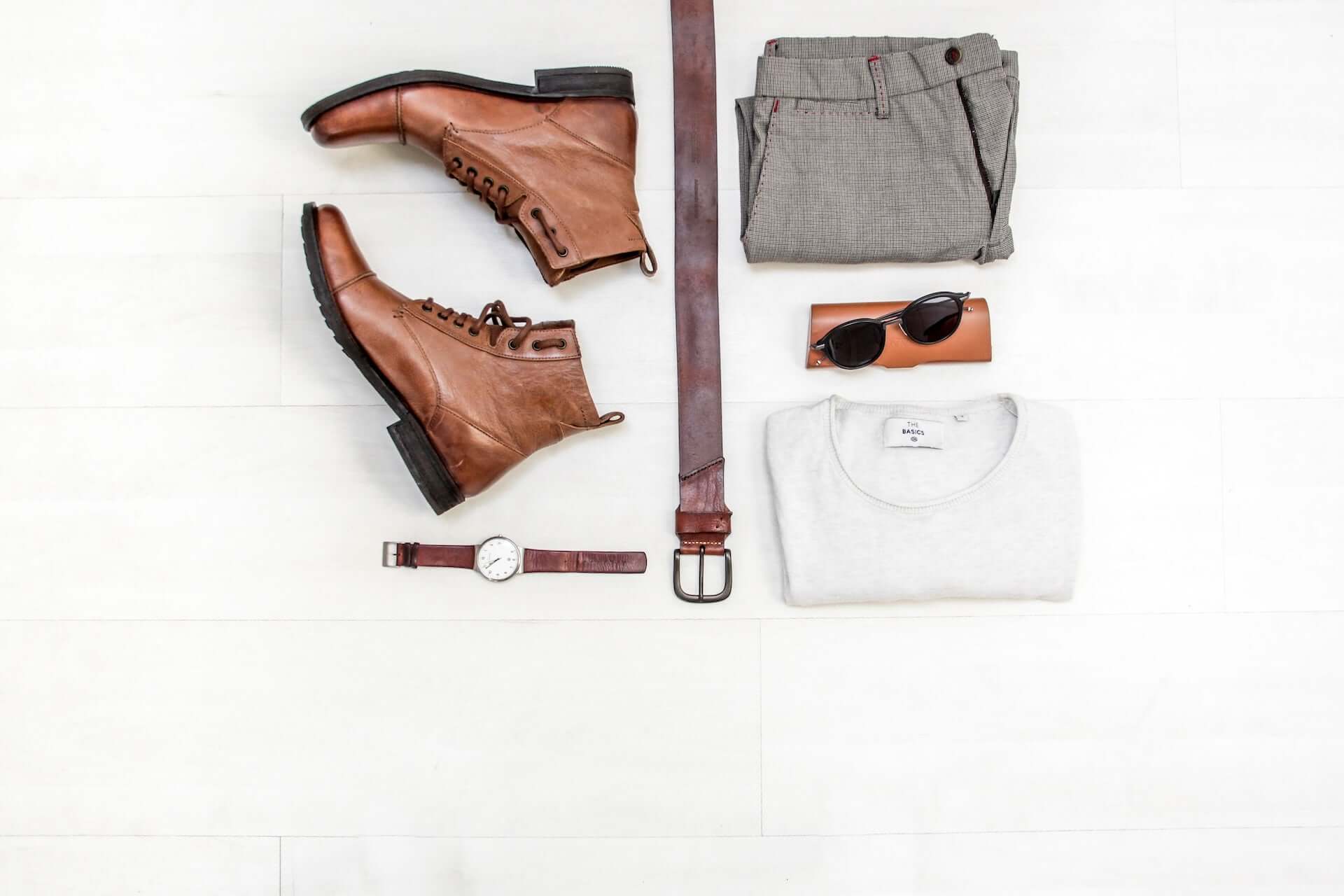 A flatlay of a simple outfit with a white shirt, grey pants, and brown accessories.