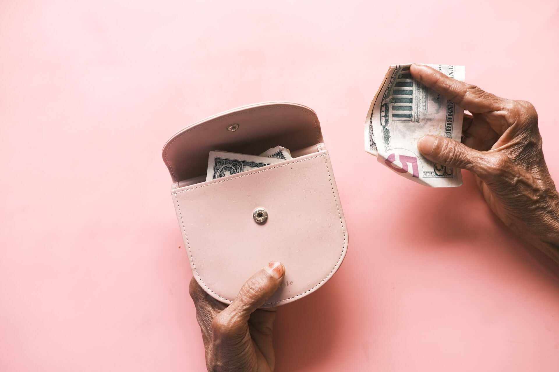 Older hands hold a small pink wallet and a five dollar bill.