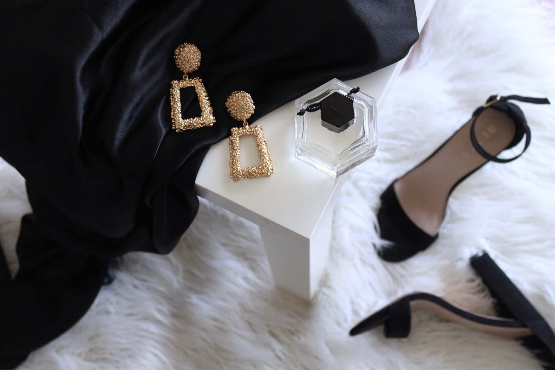 A black dress and heels with gold earrings and a bottle of perfume on a white fur rug and white minimalist table.
