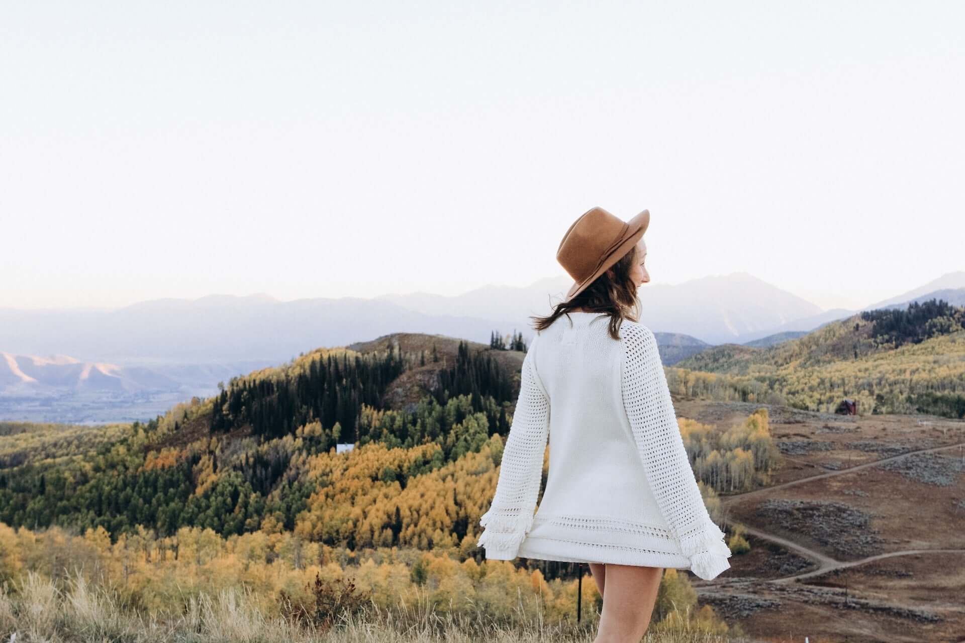 A woman in a white sweater and brown leather hat stands back to the camera in front of a forested hillside in the autumn.