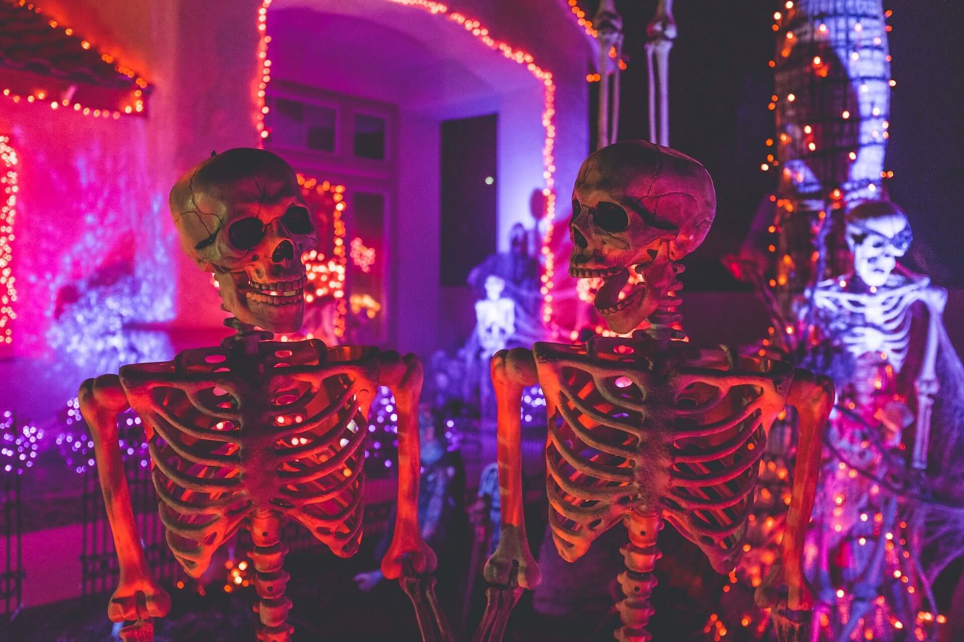 Two skeletons lit by pink neon lights in front of a decorated porch.
