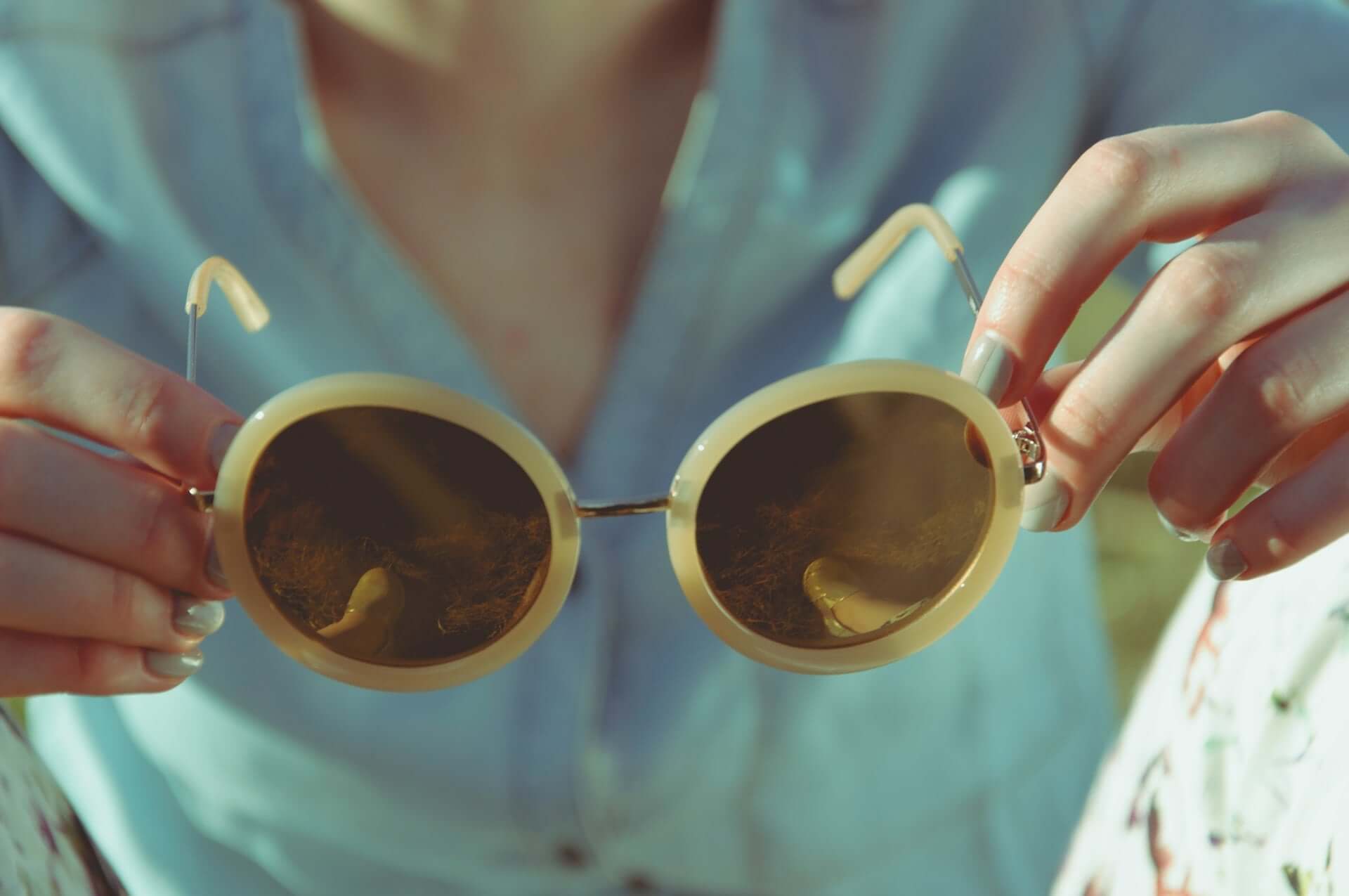 A woman in a blue button down top holds out round vintage sunglasses with off-white frames.