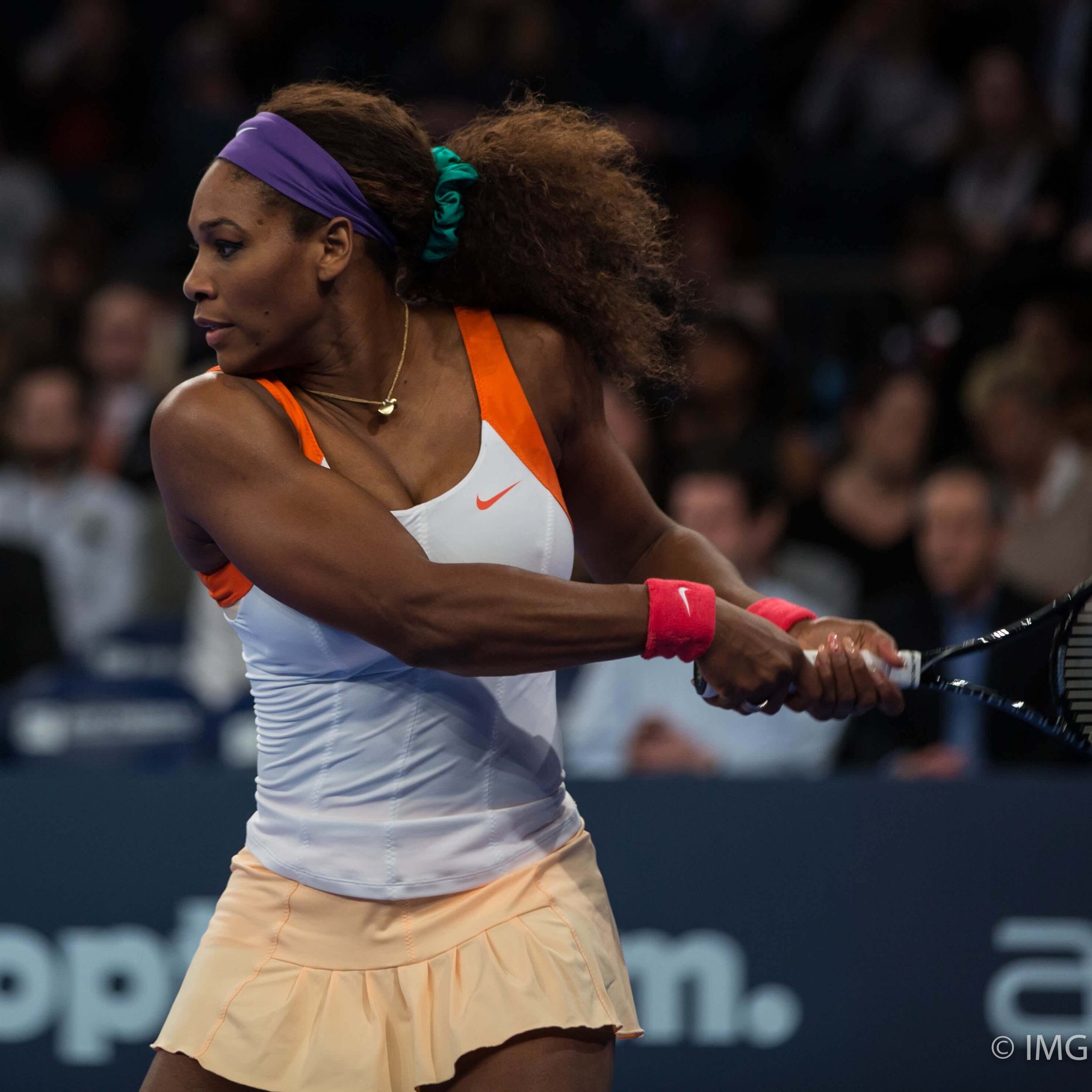 Serena Williams on the tennis court, mid swing. She is a black woman with long hair held back in a loose ponytail by a blue scrunchie. She wears a white and orange tennis tank and shirt. 