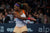 Serena Williams on the tennis court, mid swing. She is a black woman with long hair held back in a loose ponytail by a blue scrunchie. She wears a white and orange tennis tank and shirt. 