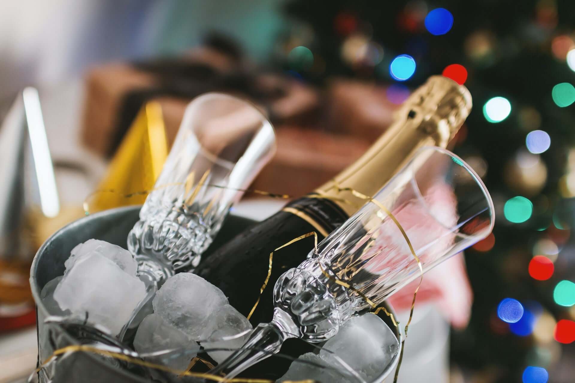 A bottle of sparkling wine and two glasses in a bucket of ice, with fairy lights in the background.