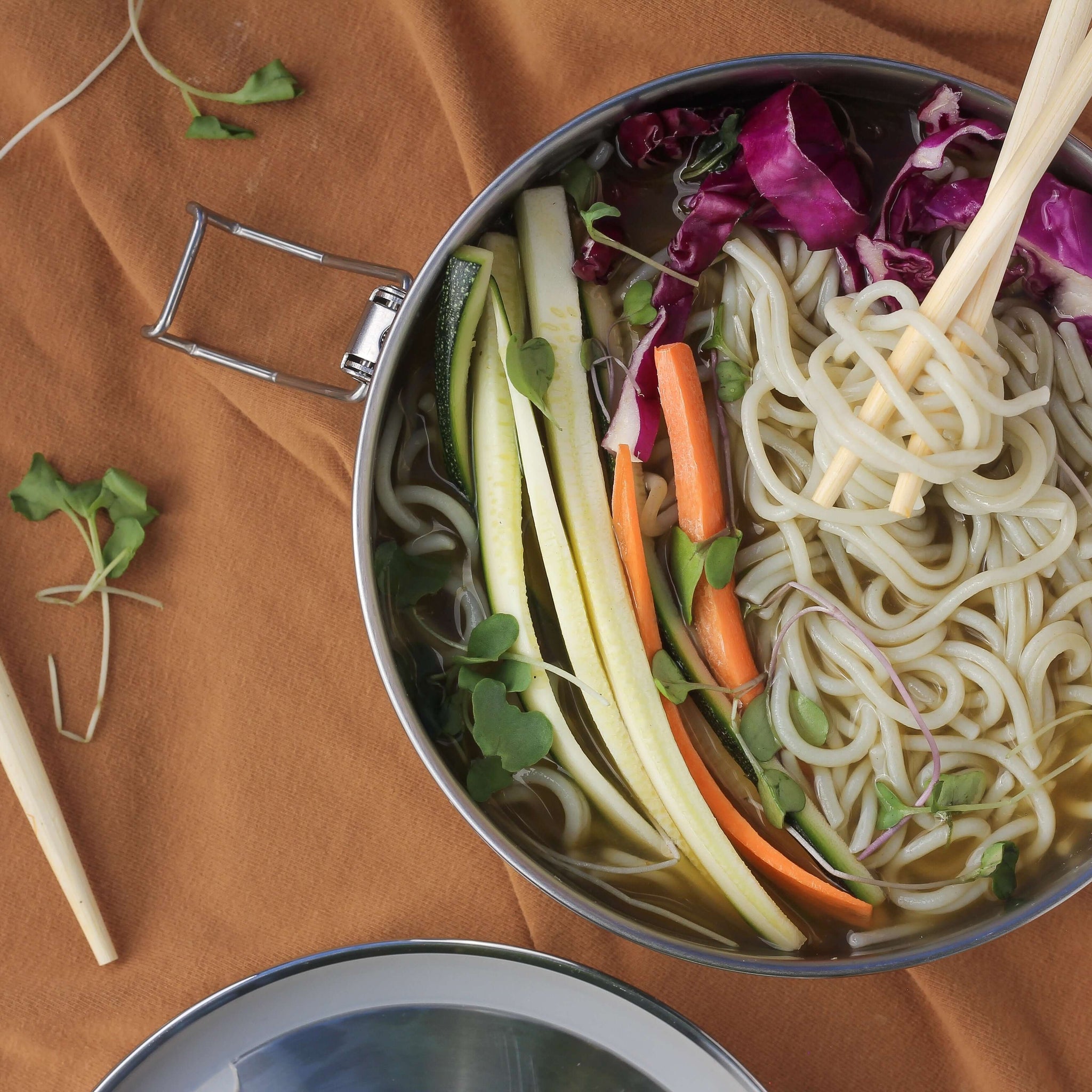 An overhead shot of a bowl full of noodles and various veggies, with chopsticks layed out beside it.