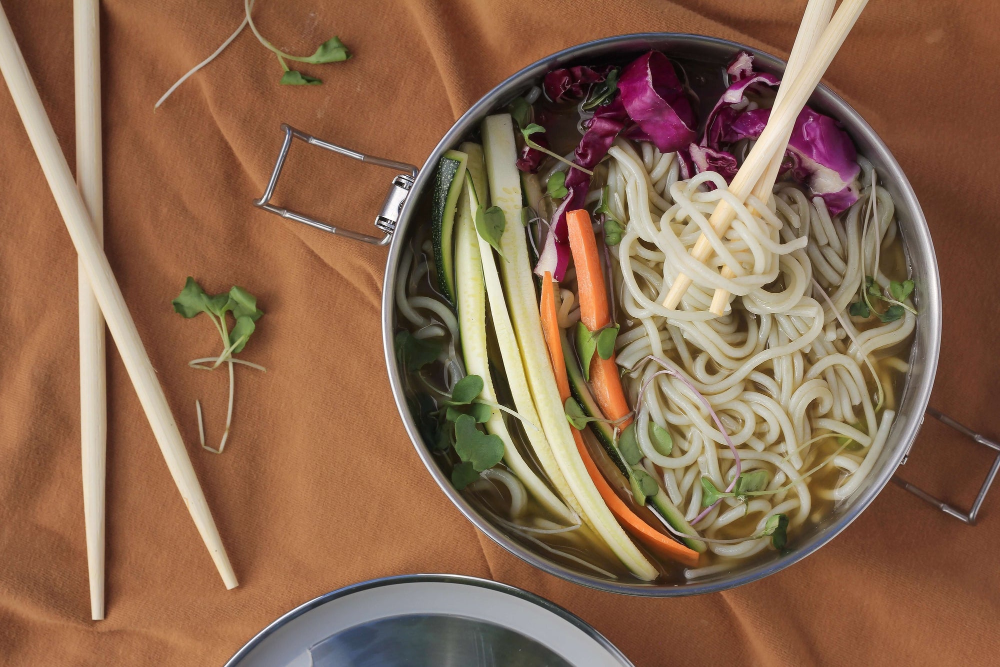 An overhead shot of a bowl full of noodles and various veggies, with chopsticks layed out beside it.