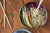 An overhead shot of a bowl full of noodles and various veggies, with chopsticks layed out beside it.