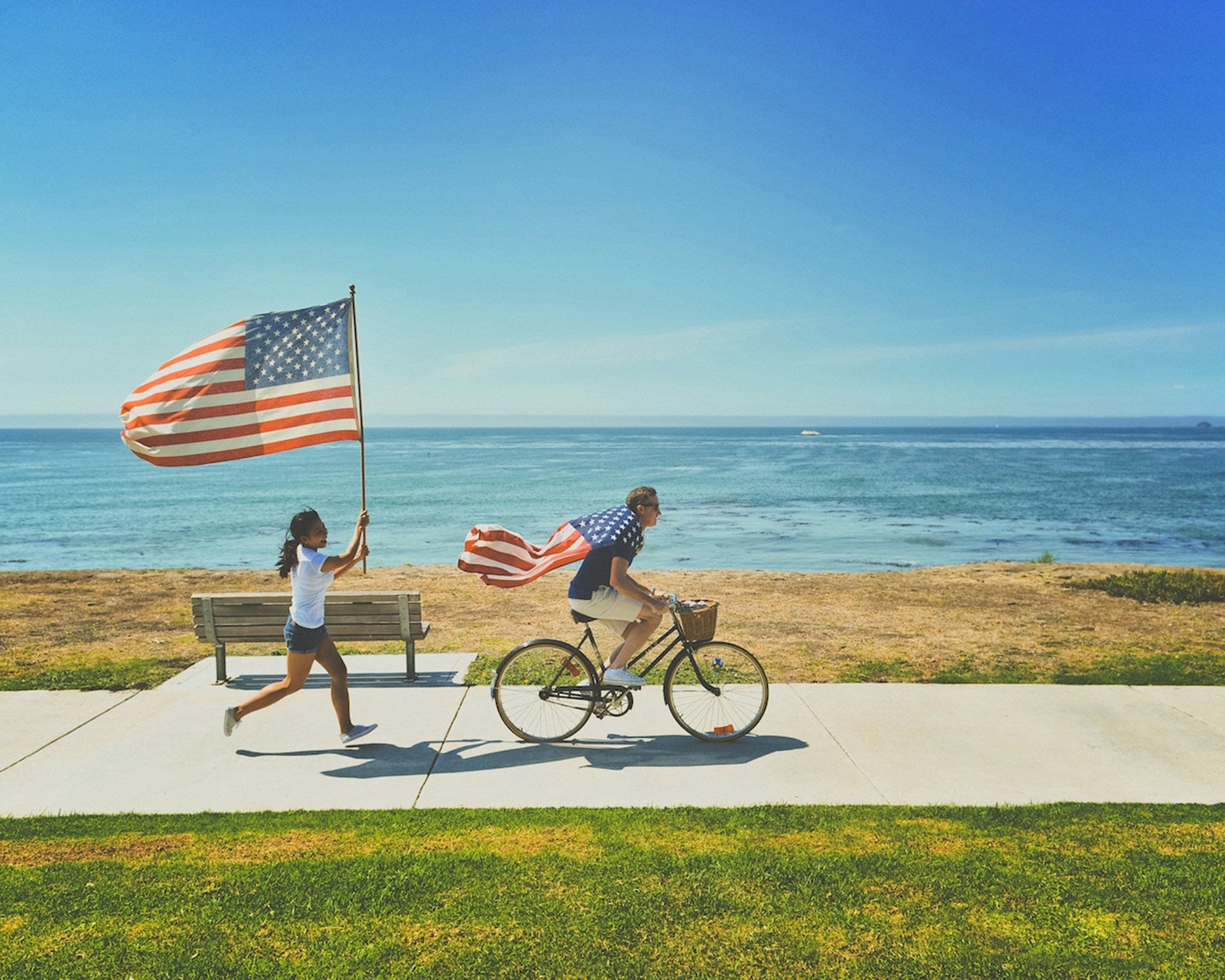 A man wearing an American flag like a cape bikes along a shorefront while a woman carries an American flag as she runs behind him.