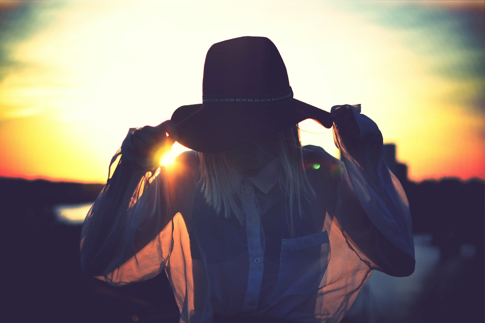 A woman backlit by the setting sun, wearing a translucent button down and a hat.